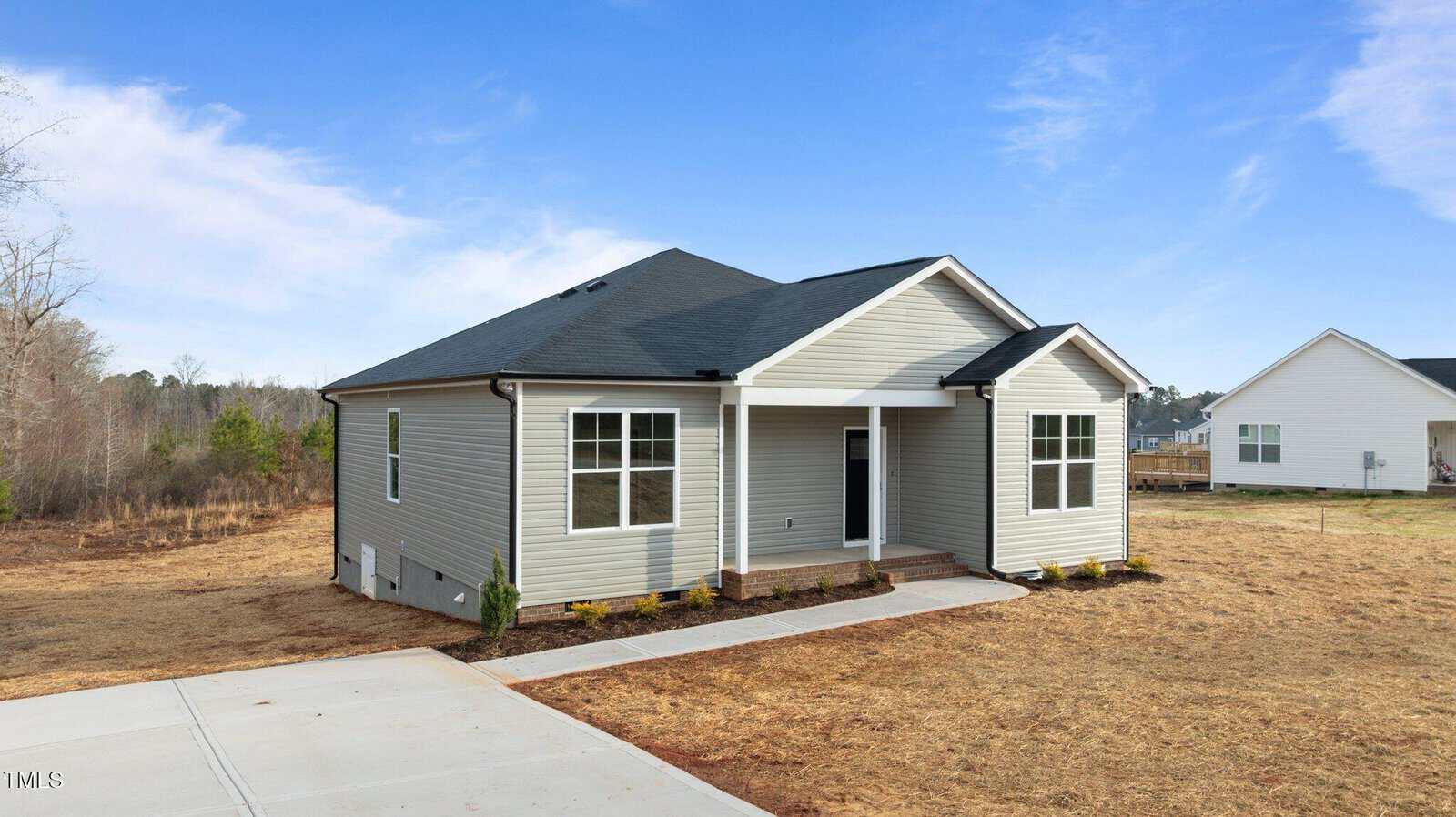 A single-story, light gray house with white trim and a covered front porch sits on a dry, sparse lawn. The driveway and sidewalk are concrete. The sky is partly cloudy and there are neighboring houses in the background.