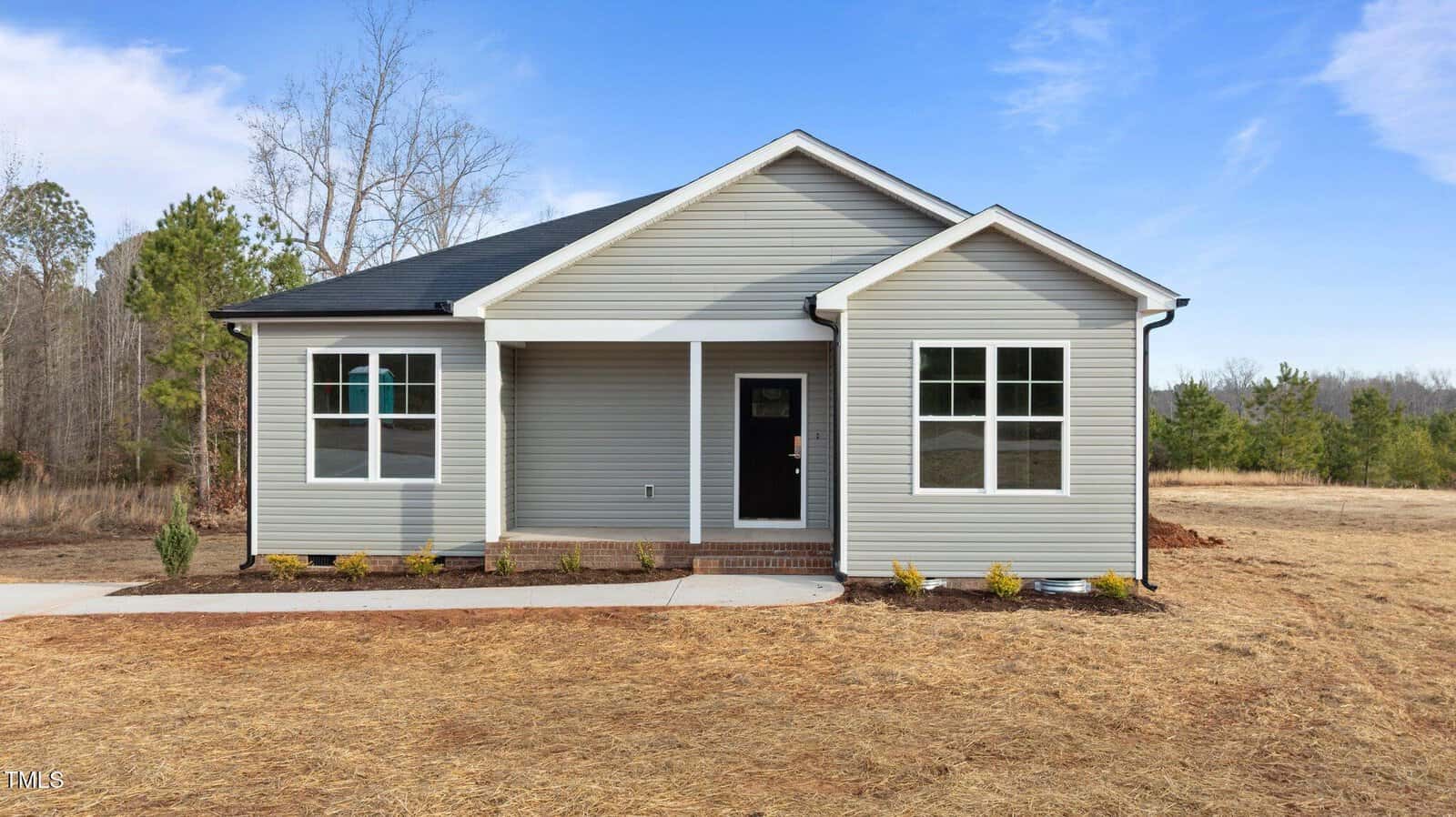 A small, single-story gray house with white trim and a black roof sits on a lot with dry grass and minimal landscaping, bordered by trees under a clear blue sky.