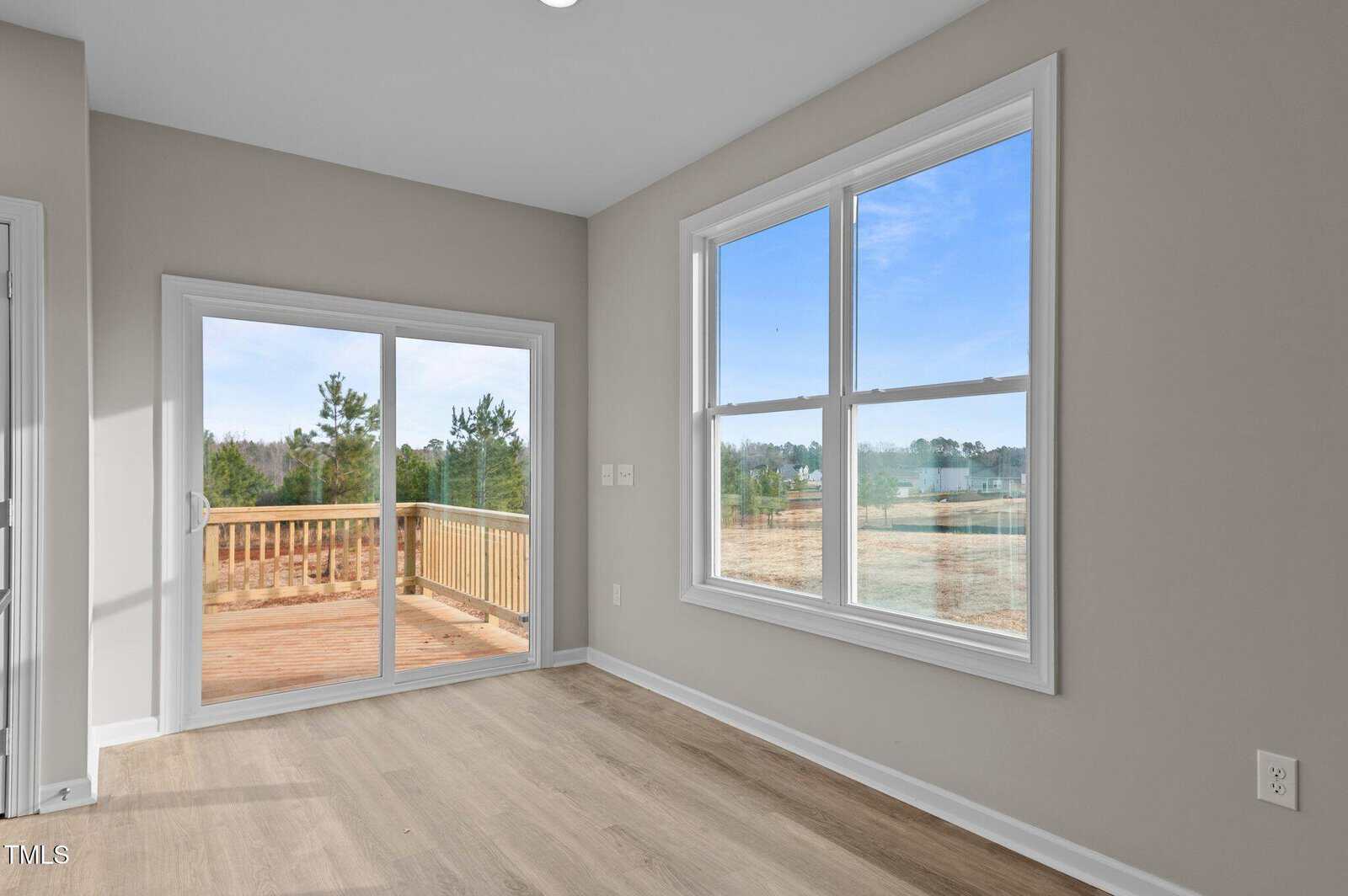 A bright, empty room with light wood flooring, light gray walls, a large double window, and sliding glass doors leading to a wooden deck with a view of trees and blue sky outside.