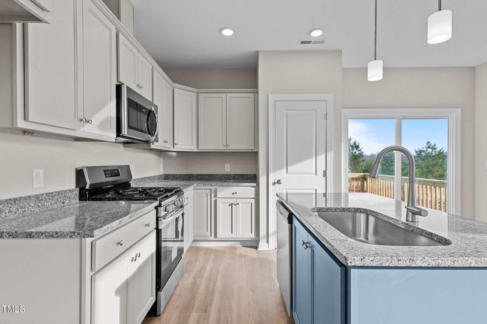 Modern kitchen with white cabinets, stainless steel appliances, granite countertops, a blue island with a sink, light wood flooring, and large sliding glass doors opening to a wooden deck with a view of trees.
