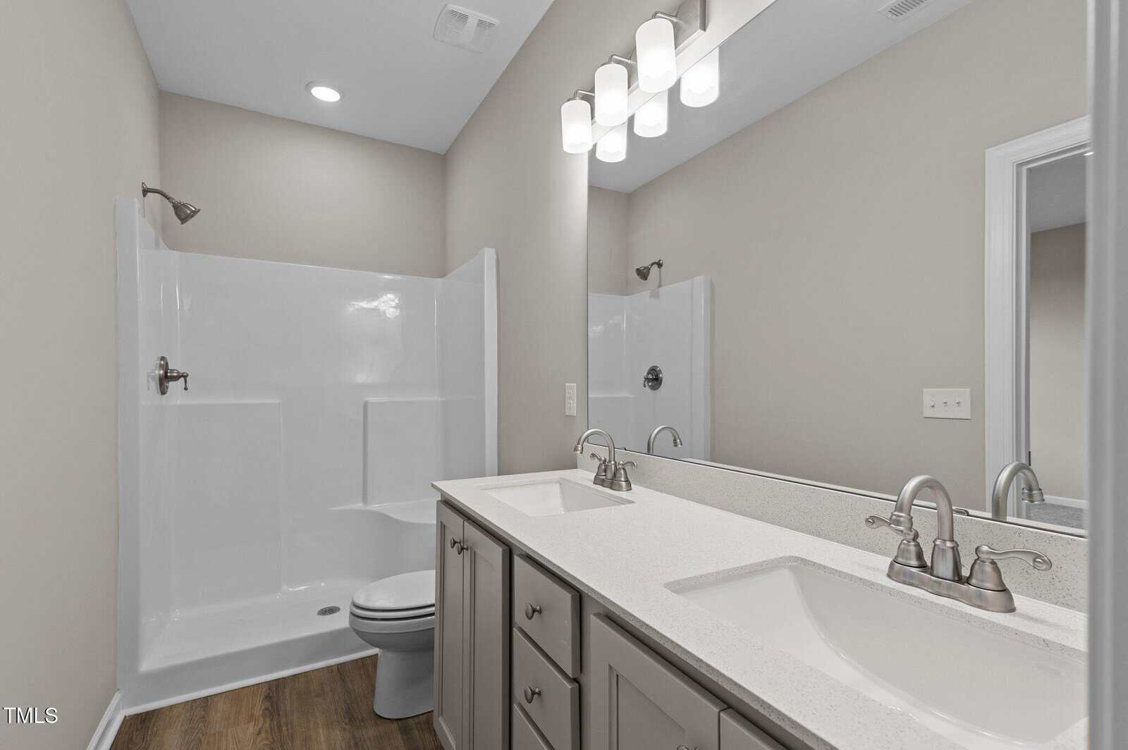 Modern bathroom with a double-sink vanity, quartz countertop, large mirror, under-mount sinks, and a walk-in shower with a built-in seat. Walls are light beige, and the floor is wood-patterned. Bright overhead lighting.