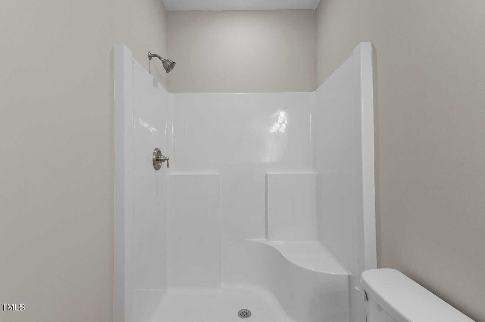 White walk-in shower with built-in seat and two wall shelves. The shower has a chrome showerhead and faucet, and is surrounded by beige walls. The edge of a white toilet is visible in the foreground.