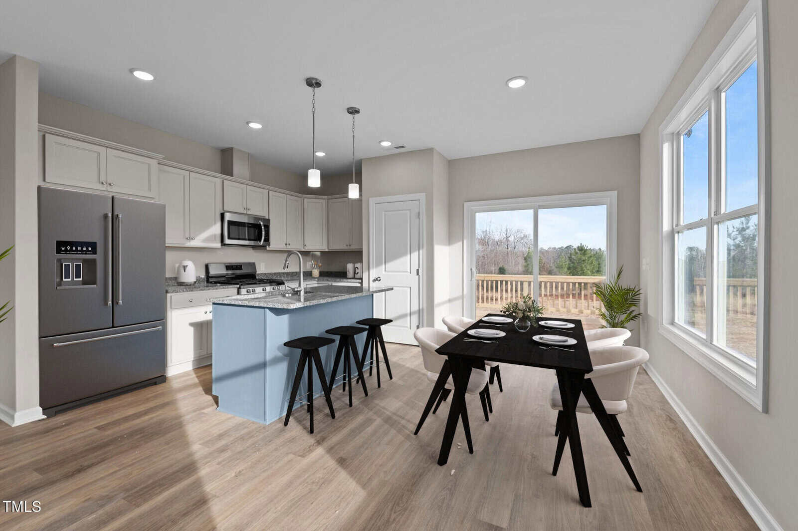 A modern kitchen with white cabinets, stainless steel appliances, a light blue island with black stools, and an adjacent dining area with a black table and white chairs next to large windows showing an outdoor deck.