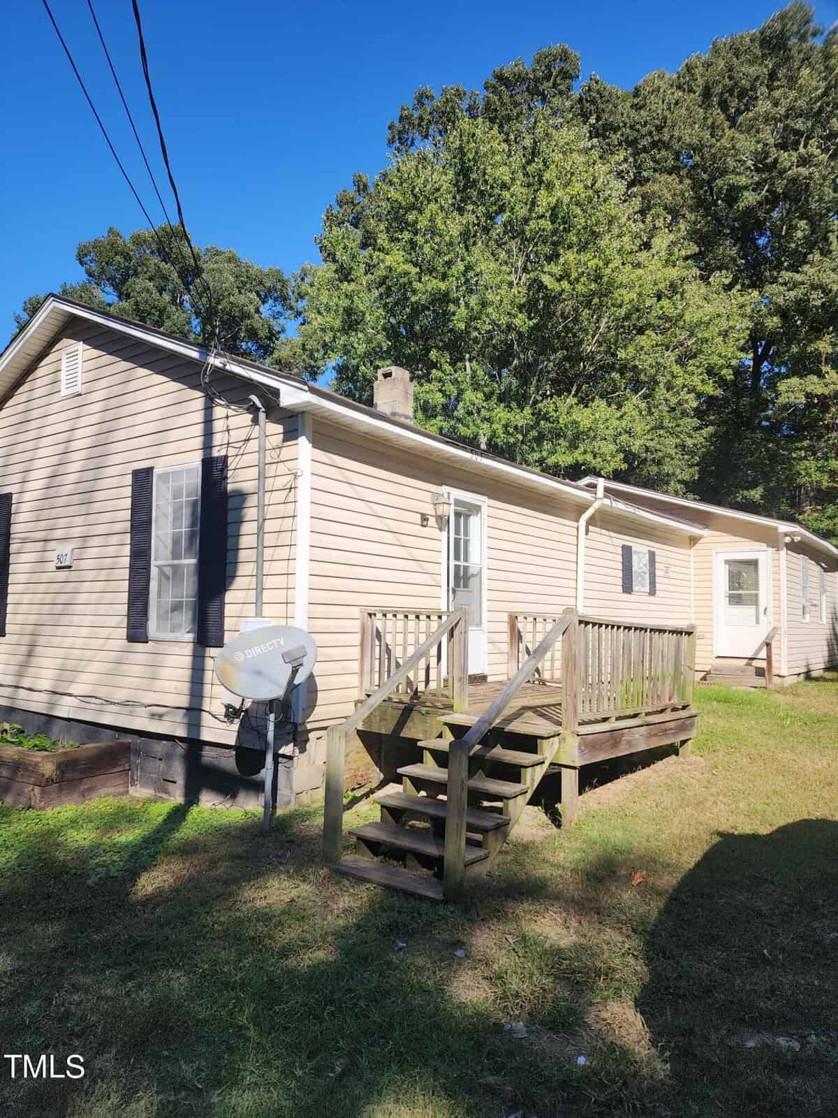 Located at 507 Neathery Street in Henderson, this beige single-story house features a charming wooden porch and sits in a grassy yard surrounded by trees. A satellite dish adorns the house under a clear, sunny sky.