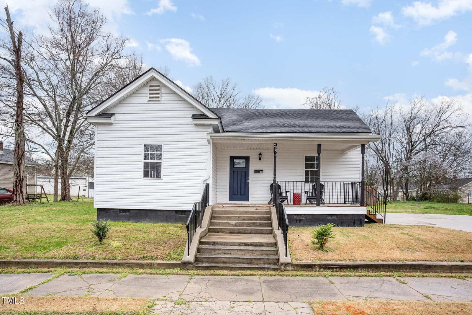 A white single-story house with a black roof, front steps leading to a blue door, black railings, a covered porch with two chairs, and a grassy yard bordered by a sidewalk. Trees are visible in the background.