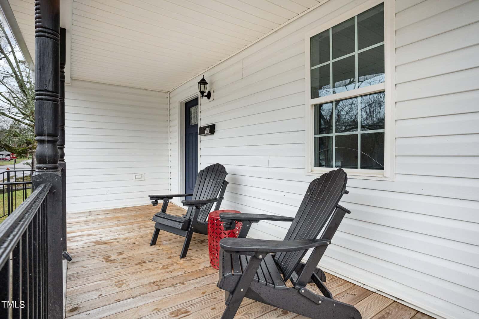 A wooden porch with two black Adirondack chairs and a small red side table. The house has white siding, a black door, a lantern-style light fixture, and a window. Trees and part of a yard are visible in the background.