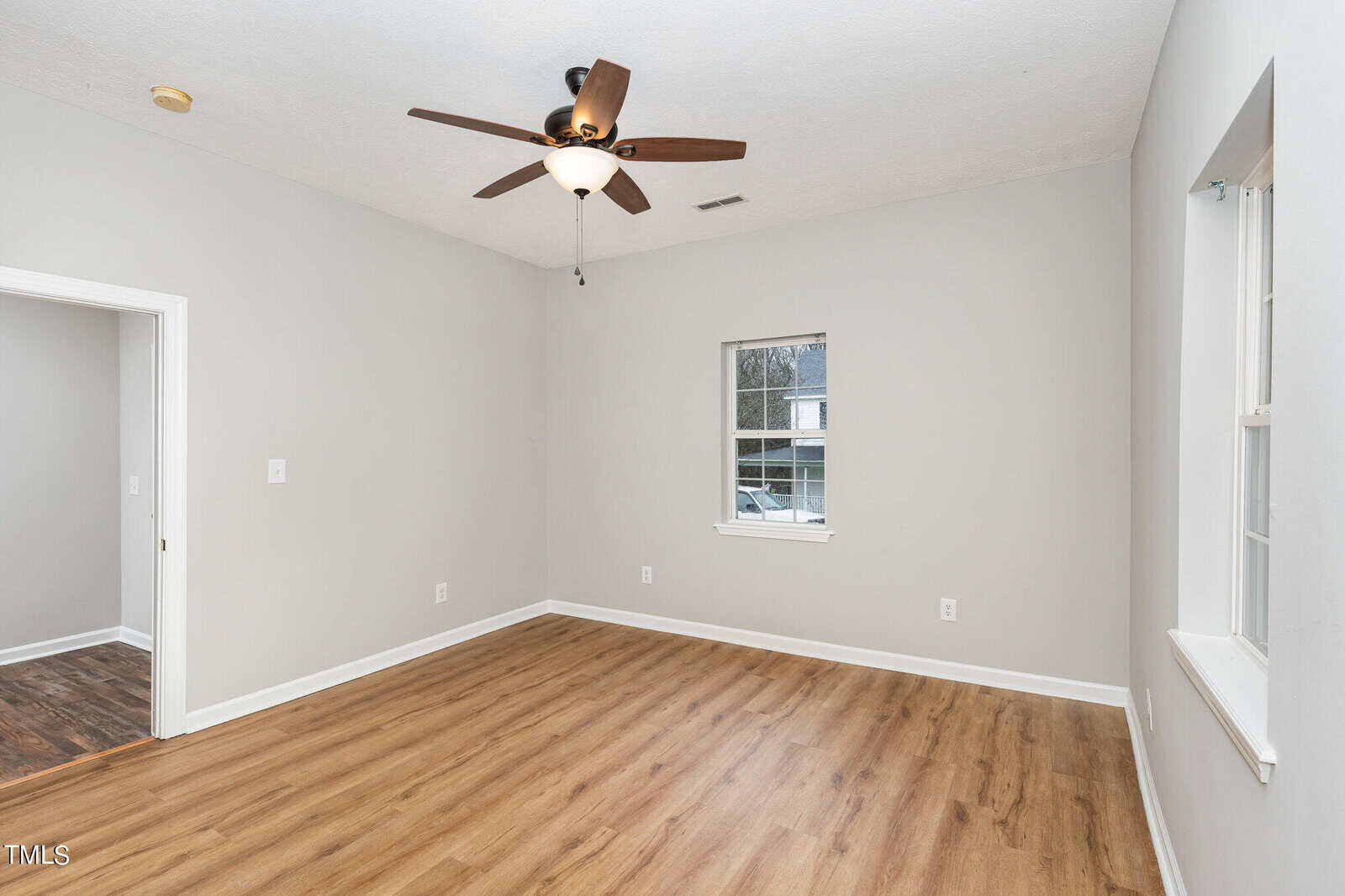 A clean, empty room with light gray walls, a ceiling fan with a light, wood-style flooring, a single window, and an open doorway leading to another area with similar flooring.