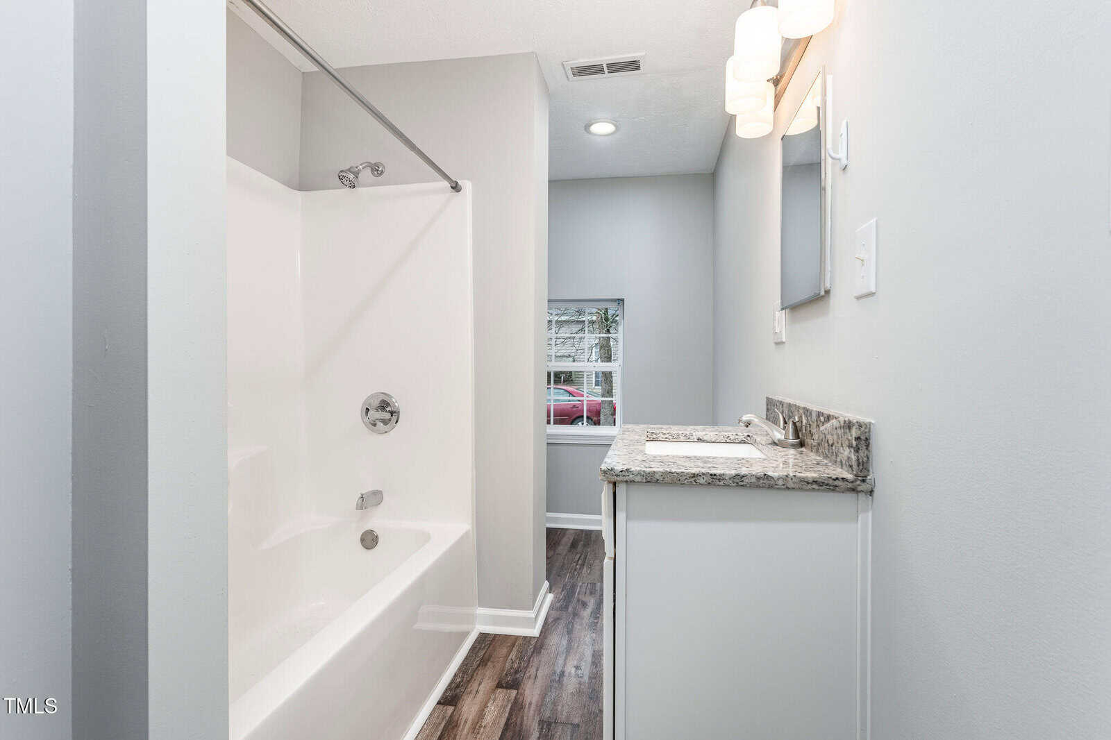 Modern bathroom with a white bathtub and shower, granite countertop vanity, large mirror, wall lights, and wood-look flooring. A window provides natural light near the back wall.