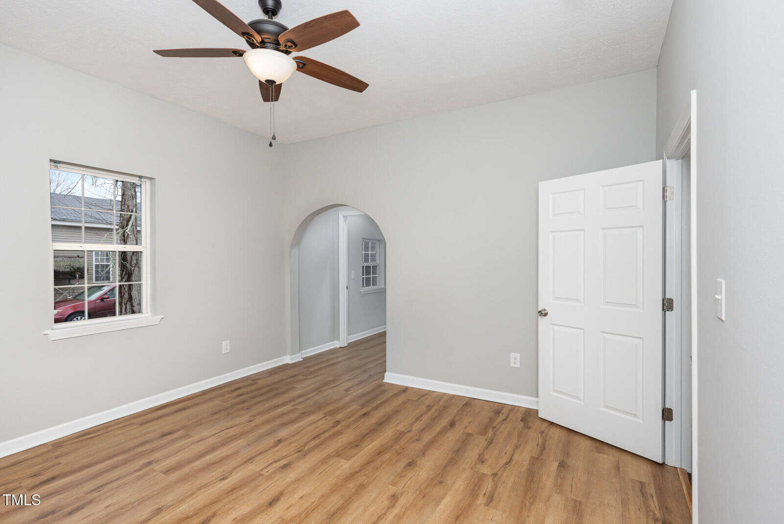 A room with light gray walls, wood-look flooring, a white door, a window, and an arched entryway leading to another room. A ceiling fan with a light is mounted on the textured ceiling.