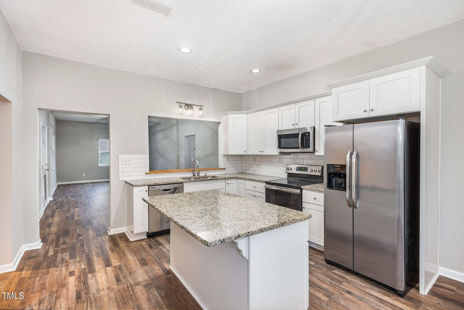 Modern kitchen with white cabinets, granite island, stainless steel appliances, and wood-look flooring. Open layout leads to another room with similar flooring and natural light. Neutral wall colors throughout.