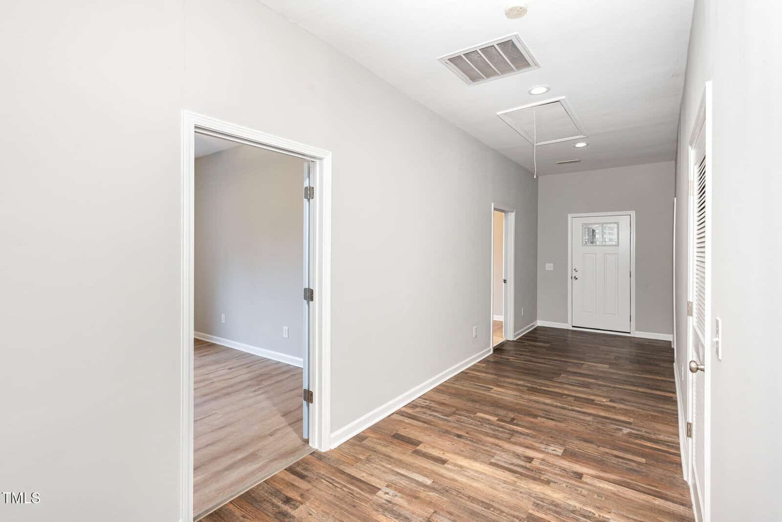 A hallway with light gray walls and dark wood flooring, leading to a white front door. An open door on the left reveals another room with similar flooring. An attic access panel is visible on the ceiling.