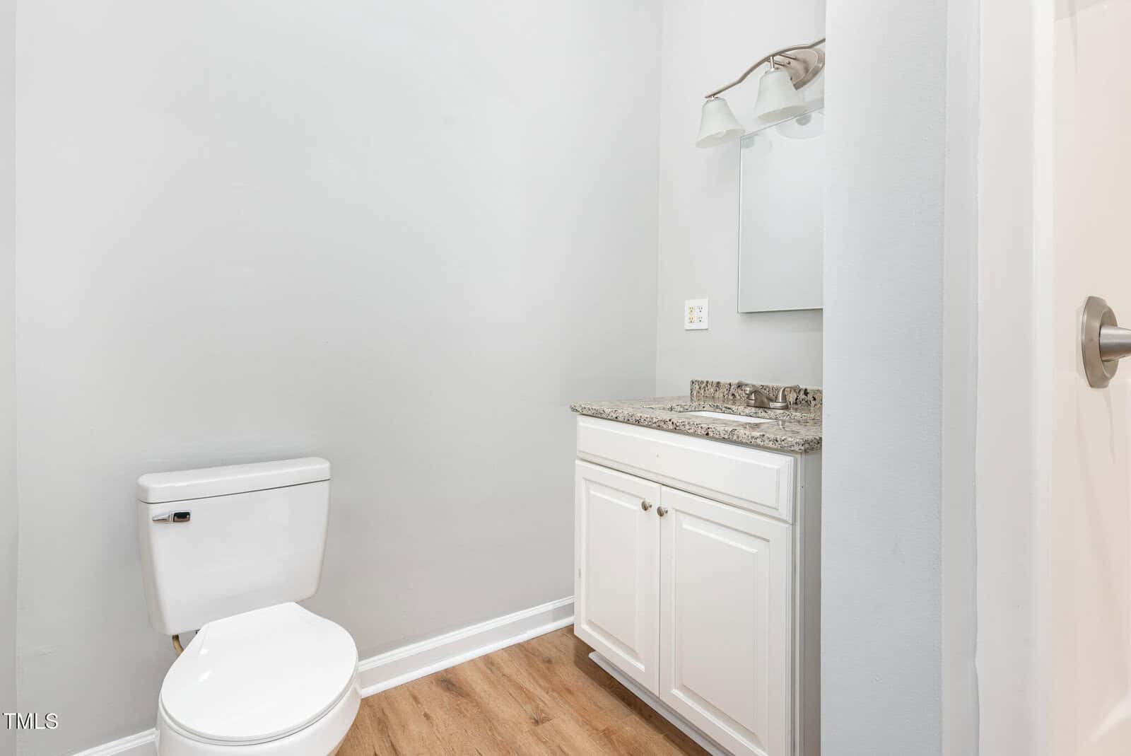 A small bathroom with a white toilet, a white vanity with a granite countertop, a wall-mounted mirror, and a light fixture above the mirror. The floor is wood-style, and the walls are painted light gray.