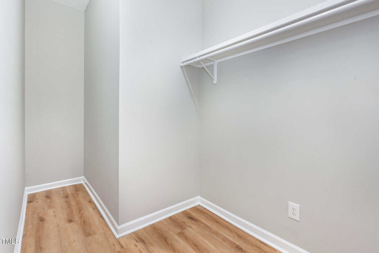 Empty walk-in closet with light gray walls, a wooden floor, a single white shelf with a hanging rod, and an electrical outlet on the right wall.