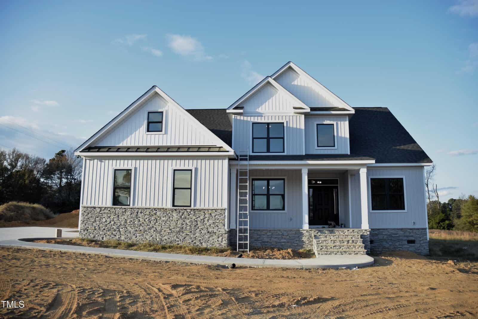 Nestled in Franklinton, the newly constructed two-story house at 4301 Bragg Farm Lane showcases white siding and a dark roof. Its stone facade graces the lower half, while a ladder leans against the front. The front yard is unlandscaped with bare dirt and a paved driveway framed by trees in the background.