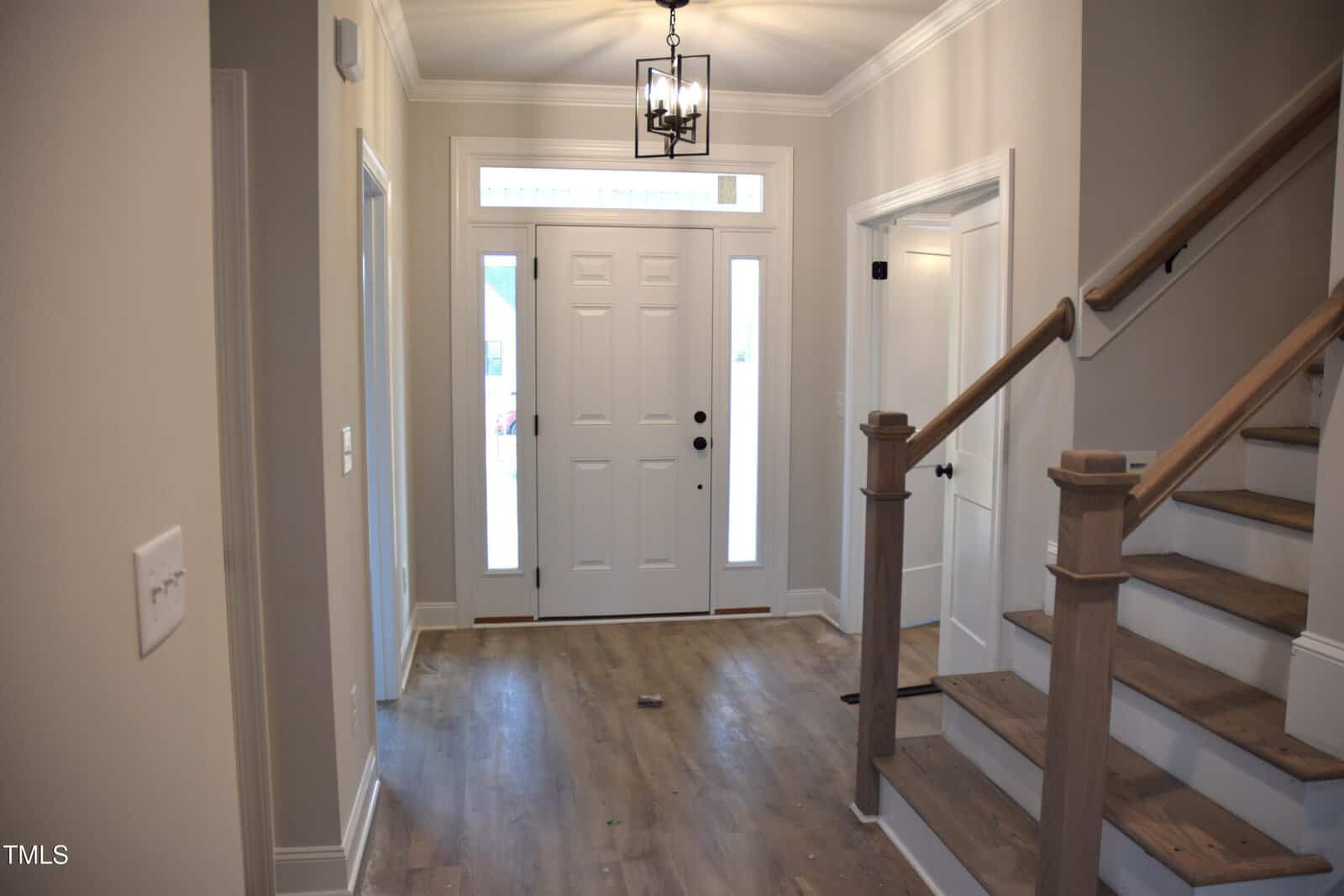 A foyer with light wood flooring and neutral-colored walls features a white front door with sidelights. A black lantern-style light fixture hangs from the ceiling. Stairs with wooden railings ascend on the right side.