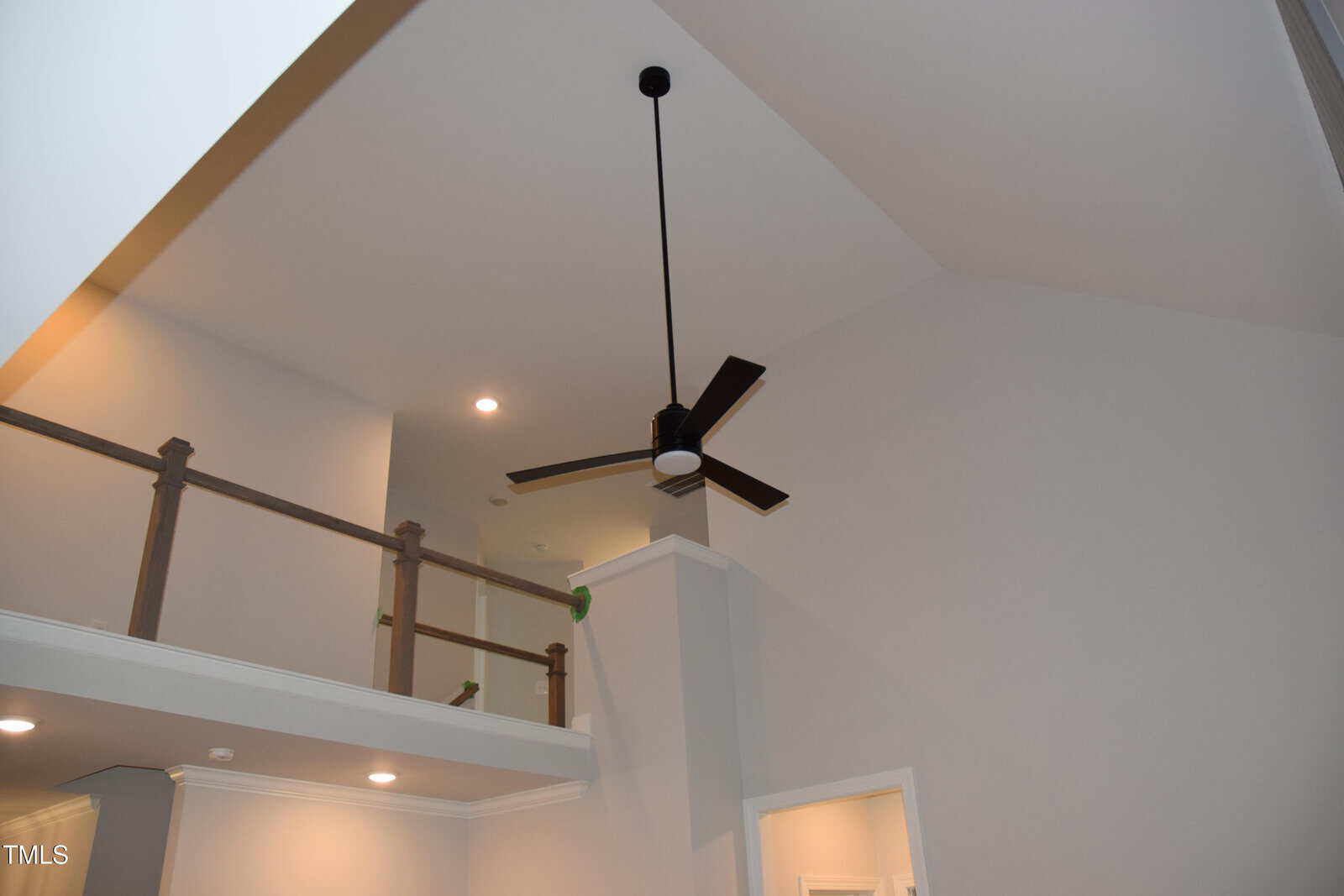 A modern ceiling fan with a circular light fixture is mounted on a high vaulted ceiling. The walls are painted light gray, and recessed lights are embedded in the ceiling. A wooden railing runs along an upper ledge.