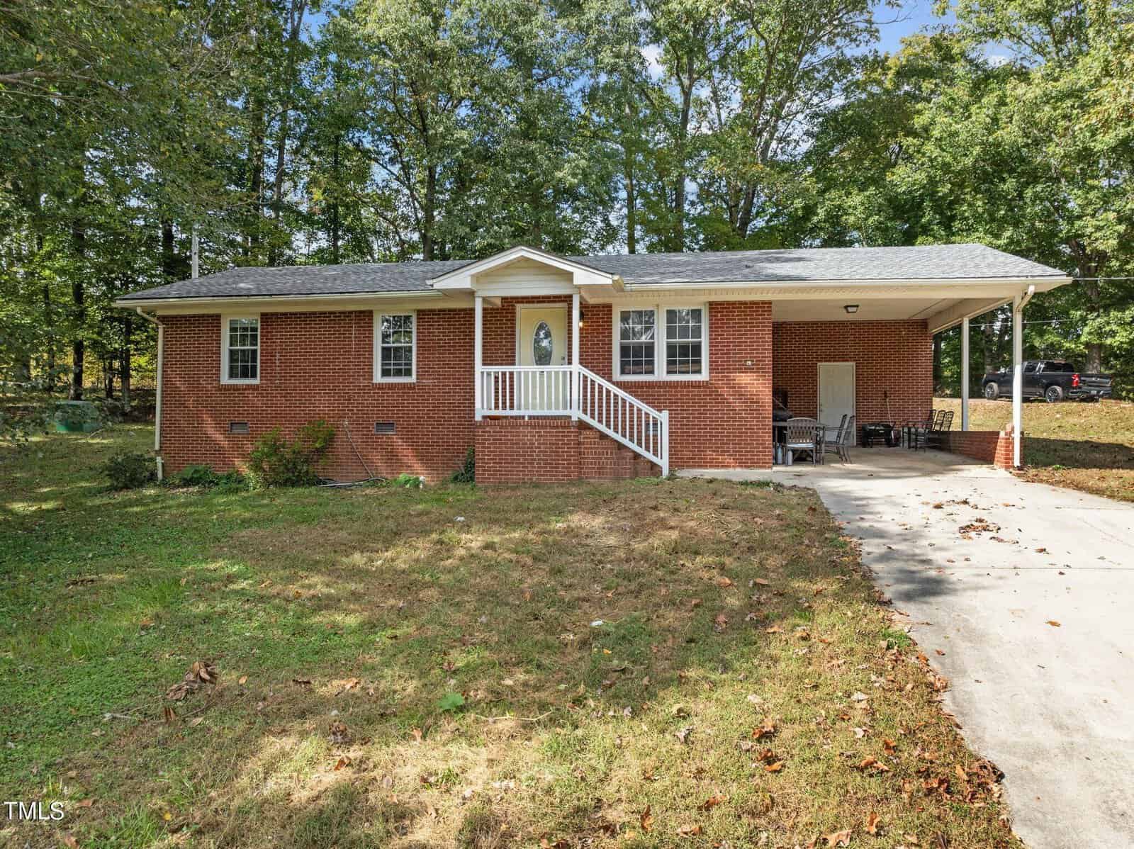 Single-story red brick house with a white front door and railing. A sloped driveway leads to a carport on the right. A few patio chairs are under the carport, and the property is surrounded by trees and grass.