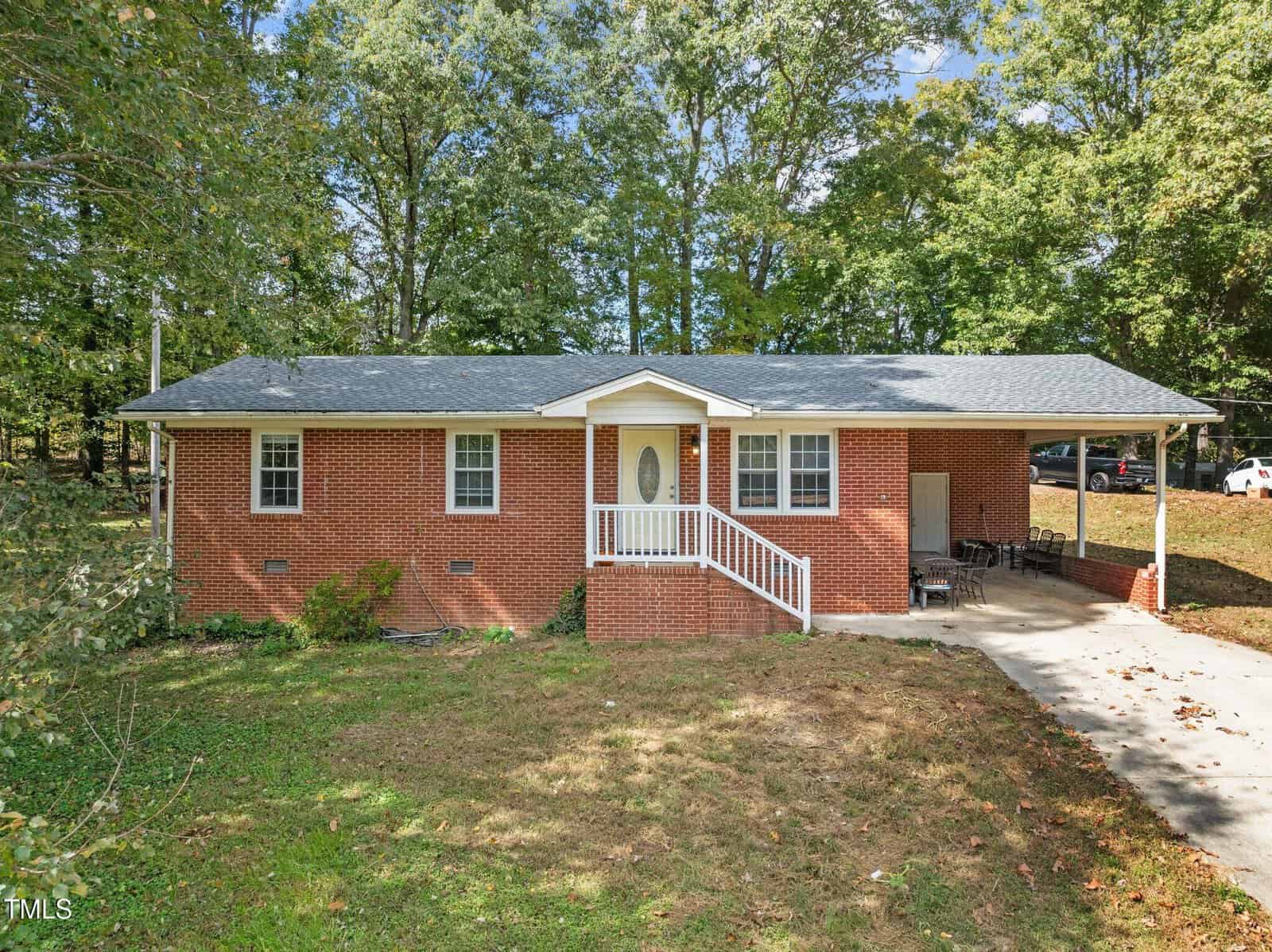 A charming single-story red brick house on 820 Egypt Mountain Road in Kittrell, featuring a cozy front porch with white railings. The gray shingle roof complements the lush green trees, while a concrete driveway leads to an attached carport on the right.