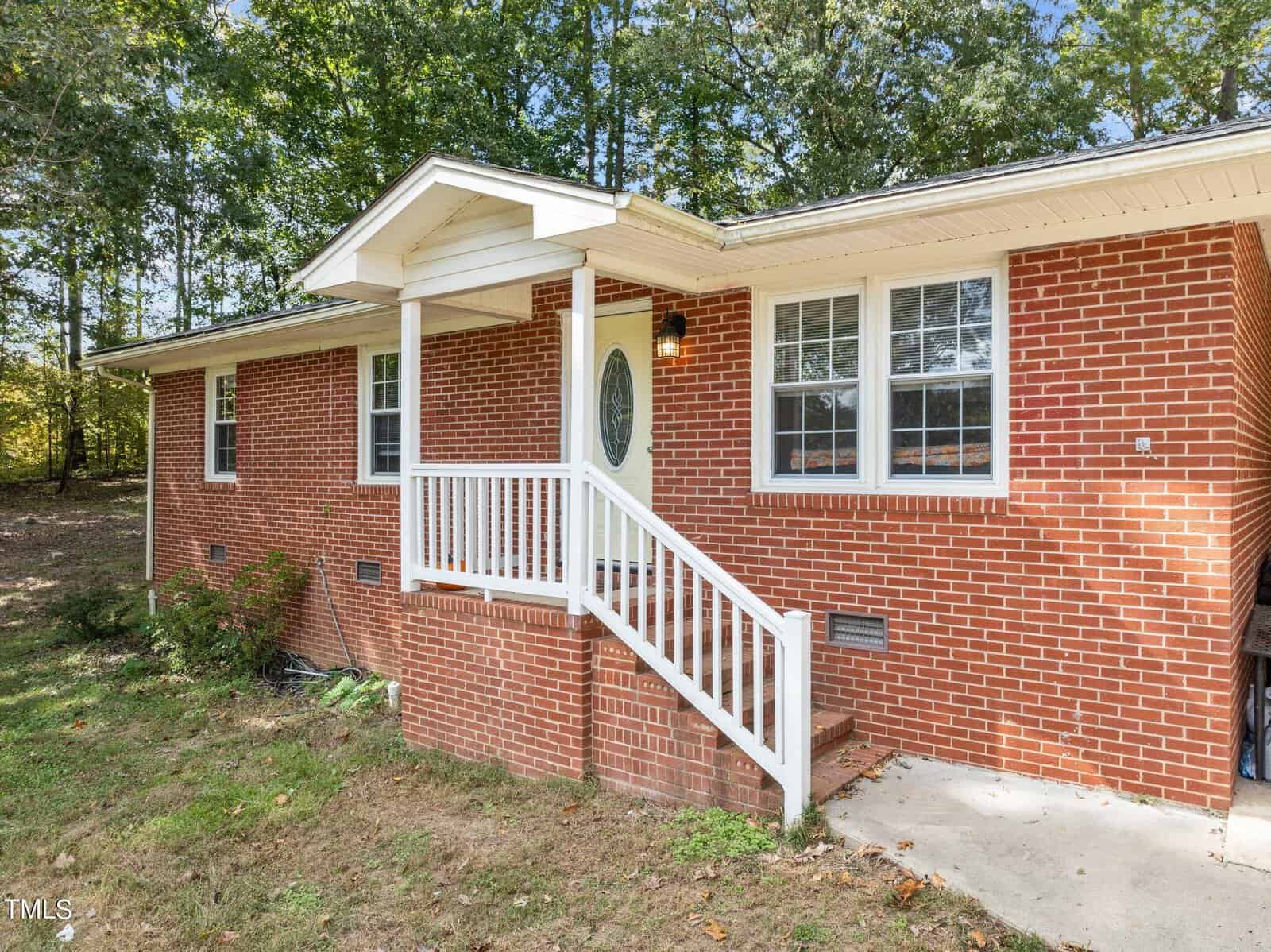 A brick house with a small white porch and steps leading to the entrance. The house has two windows with white shutters and is surrounded by a grassy area with trees in the background.
