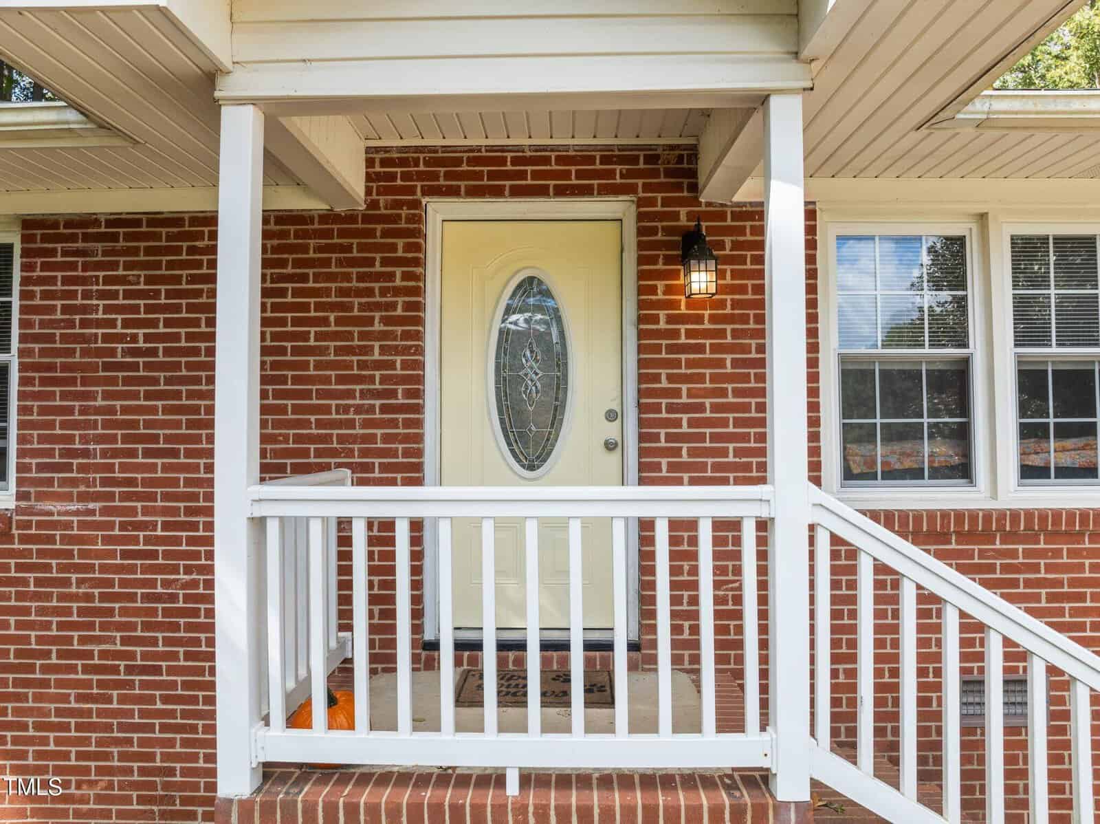 A brick house entrance featuring a white door with an oval window, surrounded by a white railing. A welcome mat lies at the doorstep, and a lantern-style light fixture is mounted next to the door. The porch has a few steps leading up to it.