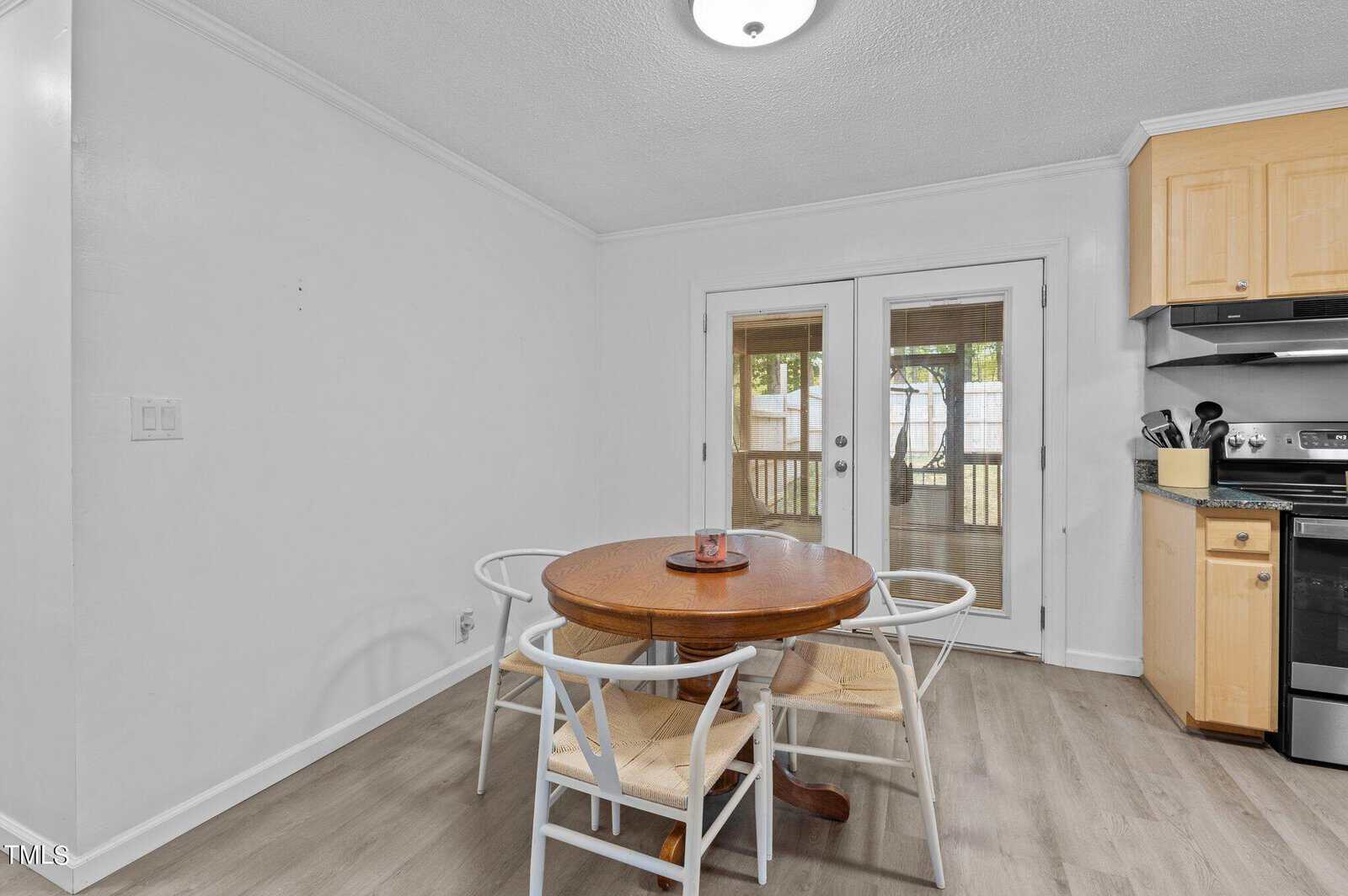 A small dining area with a round wooden table and four chairs on a light wood floor. A candle sits on the table. Double glass doors lead to an outdoor area. A white wall and part of a kitchen with light wood cabinets and a stove are visible.