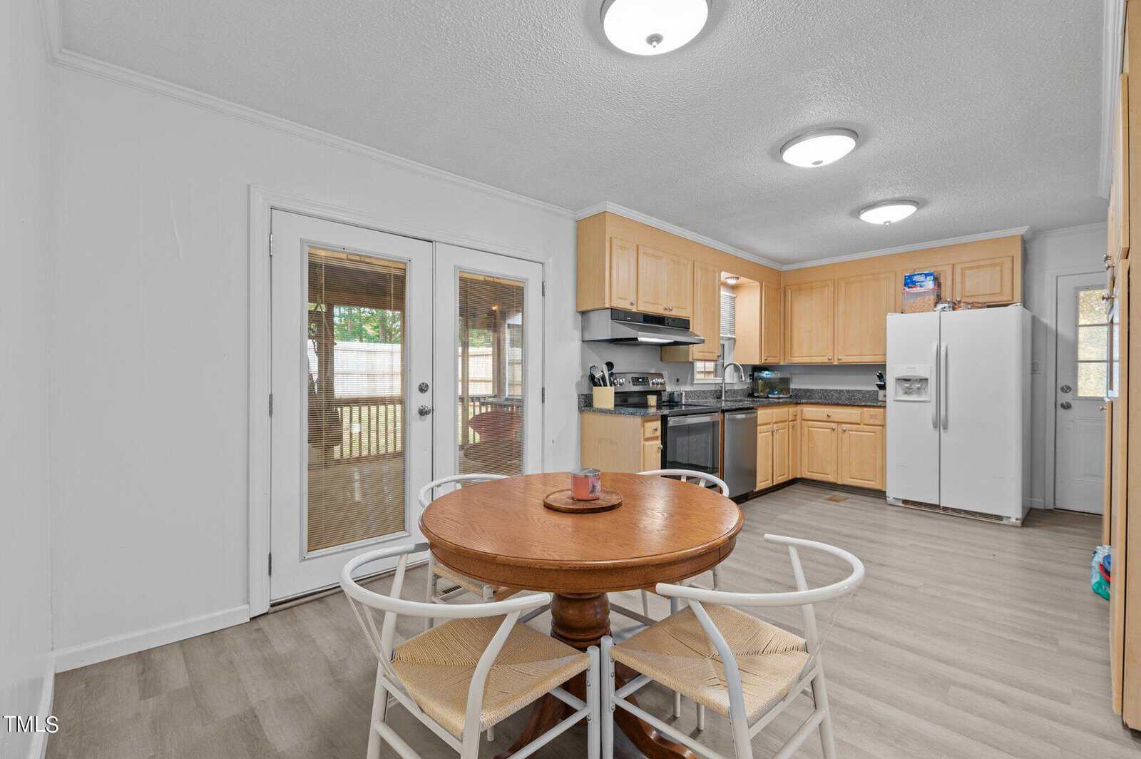 Spacious kitchen with light wood cabinets, stainless steel appliances, and a white fridge. A round wooden dining table with four chairs sits on light wood flooring. French doors open to an outdoor area, adding natural light to the space.