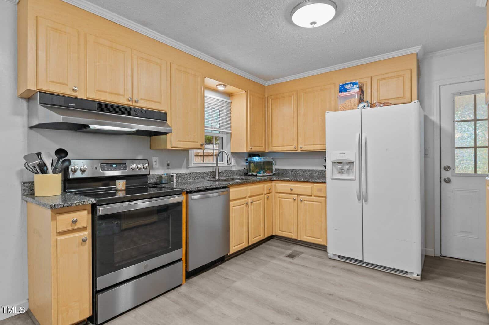 A modern kitchen featuring light wooden cabinets, a stainless steel oven, and a matching range hood. A white refrigerator stands beside the door. The countertops are gray granite, with kitchen utensils and a small fish tank. The floor is light wood.
