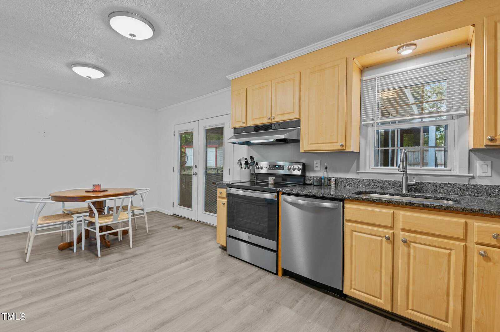 A modern kitchen features light wood cabinets, a black countertop with a stainless steel stove, and dishwasher. A small dining area with a round table and chairs is in the corner. A glass door leads to the outside, letting in natural light.