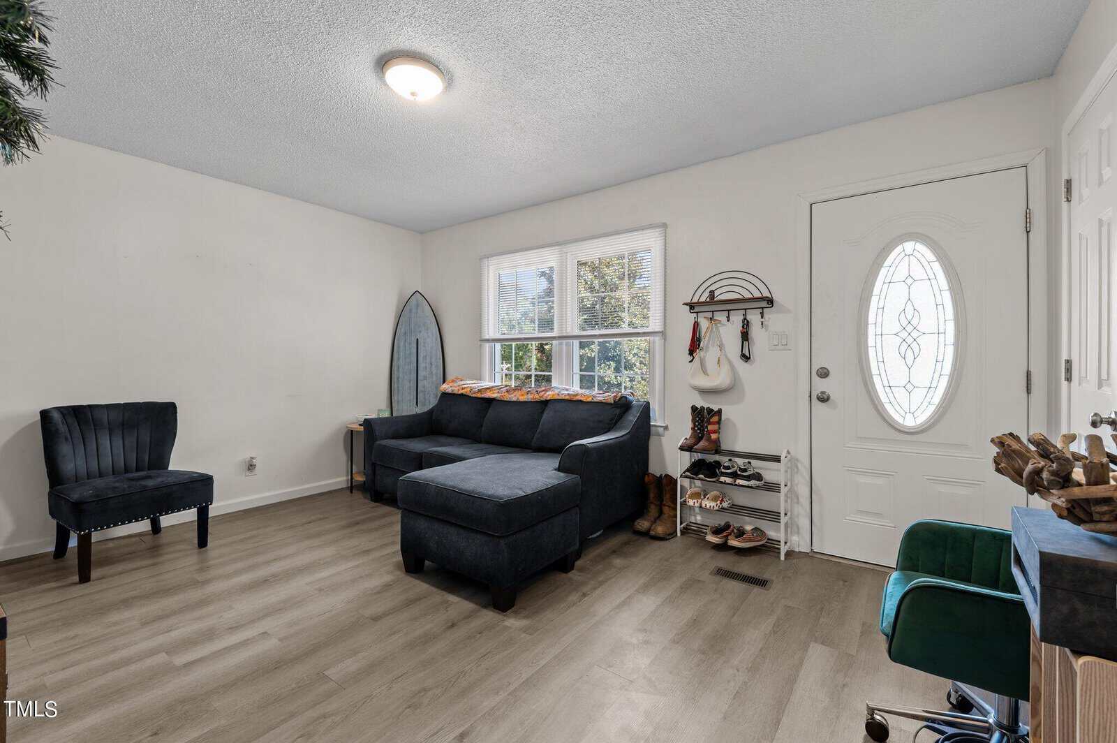 A cozy living room with a dark green sectional sofa and a matching chair. A surfboard leans against the wall. Near the door, there's a shoe rack and coat hooks. The room has light wood flooring and a round ceiling light fixture.