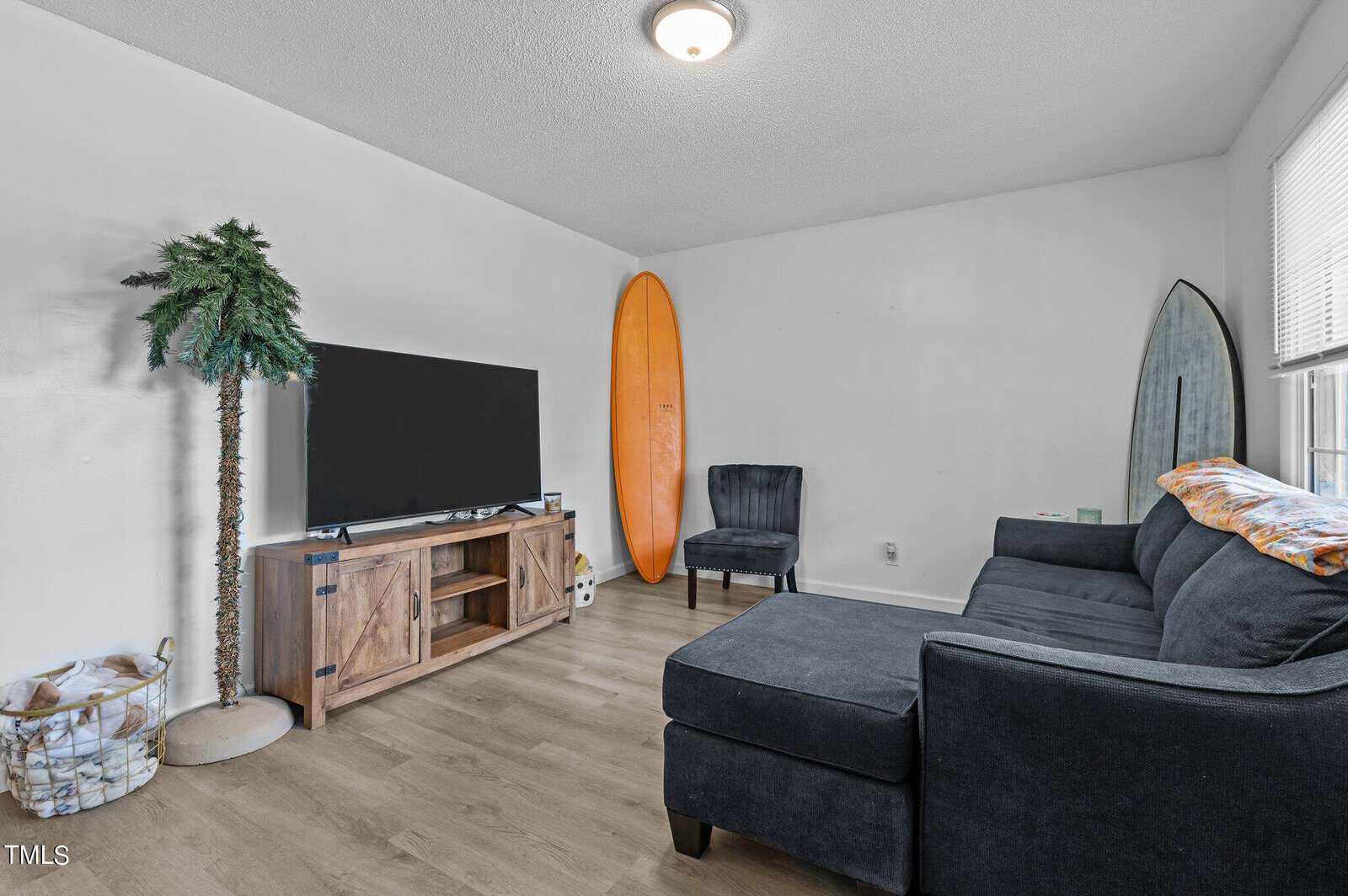 A modern living room with a black sectional sofa, a dark armchair, and a flat-screen TV on a wooden stand. Two surfboards, one orange and one mirrored, are propped against the wall. A potted palm completes the decor on light wood flooring.