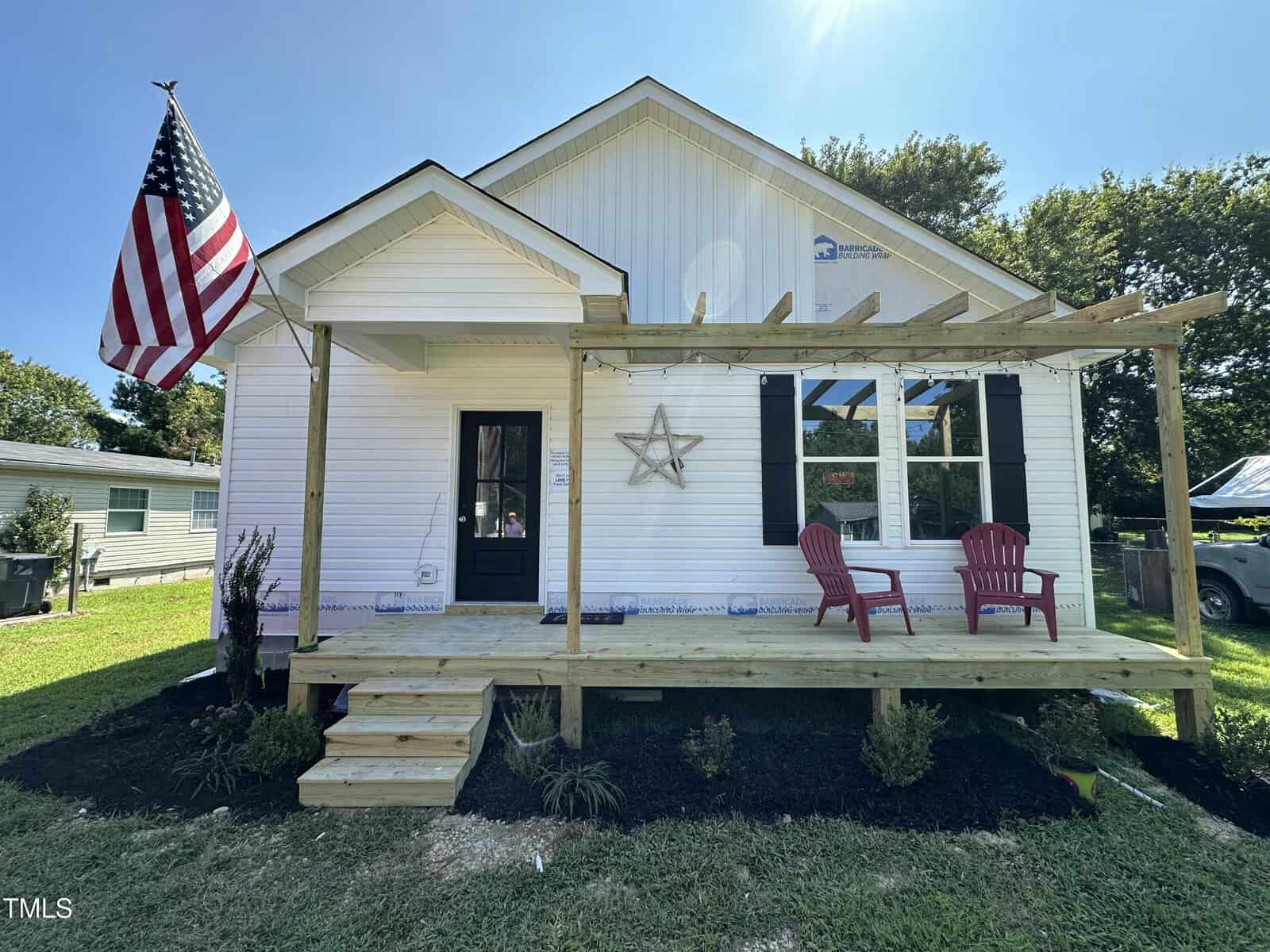 A small white house on 118 Daniel Street, with a wooden front porch, showcases an American flag, two red chairs, and a star decoration. This Oxford gem is surrounded by a neatly landscaped garden in a grassy yard framed by trees.