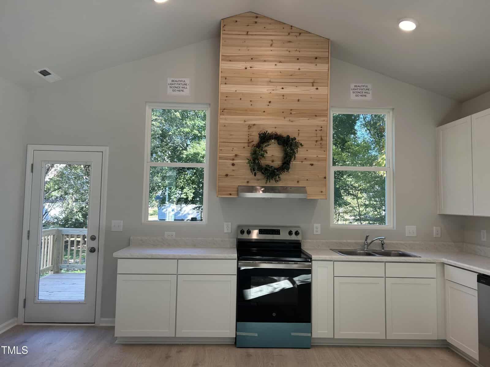 A kitchen interior with white cabinets, an electric stove, and a wooden vent hood featuring a decorative wreath. Two windows and a glass door provide natural light. Light wood flooring and an overhead light fixture complete the space.