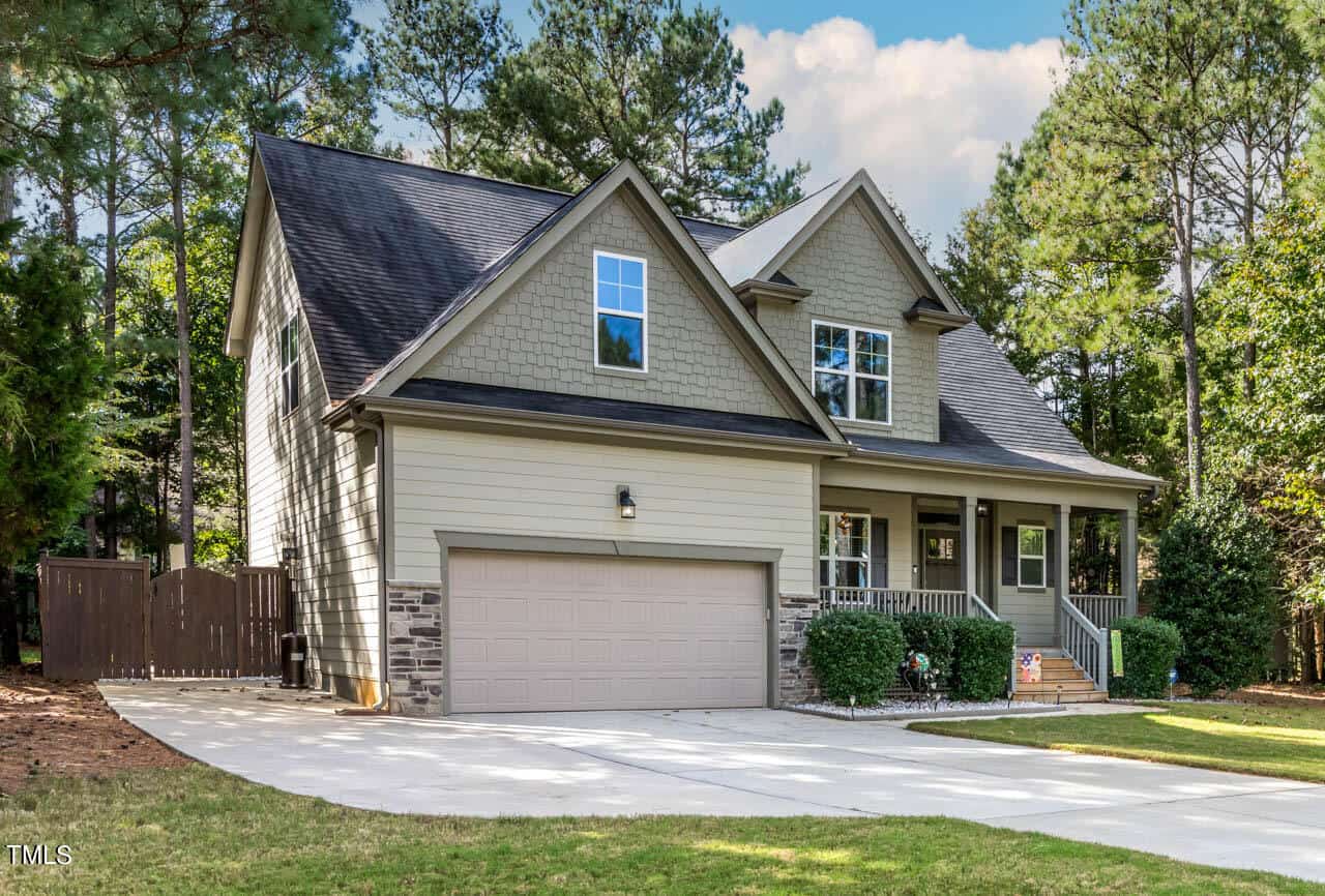 Two-story suburban house with beige siding, stone accents, a front porch, and a double garage. The home is surrounded by tall trees and greenery, with a curved driveway leading to the garage.