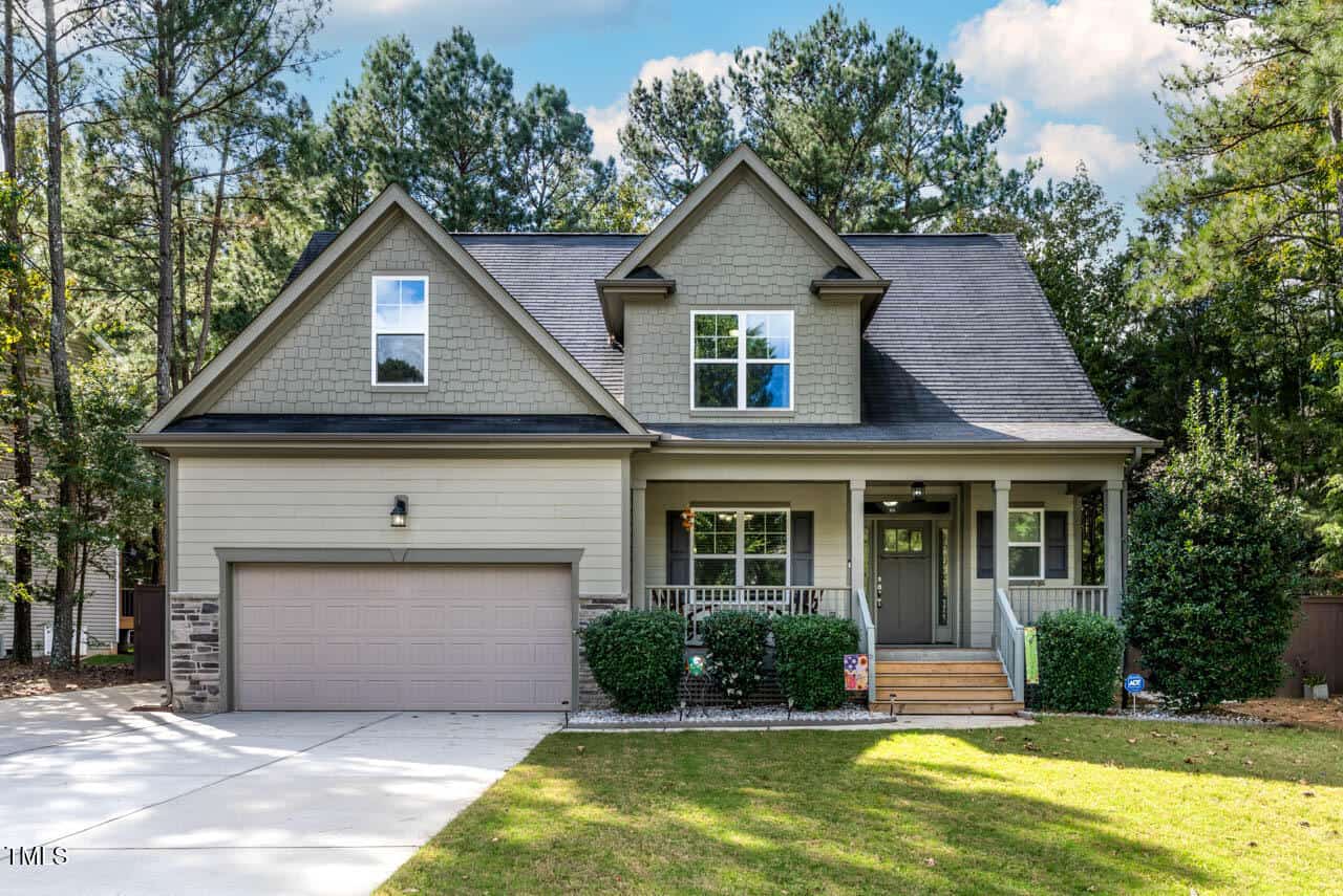 A two-story suburban house with gray siding, a front porch, a double garage, and large windows, surrounded by trees and a grassy yard under a blue sky with clouds.