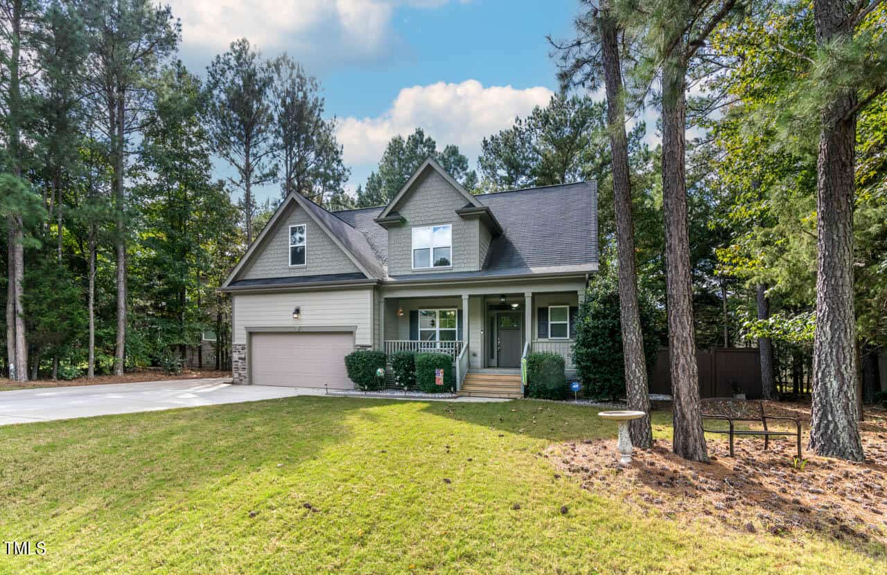 A two-story house with a gabled roof, front porch, and attached two-car garage sits on a spacious, grassy yard surrounded by tall trees under a partly cloudy sky.