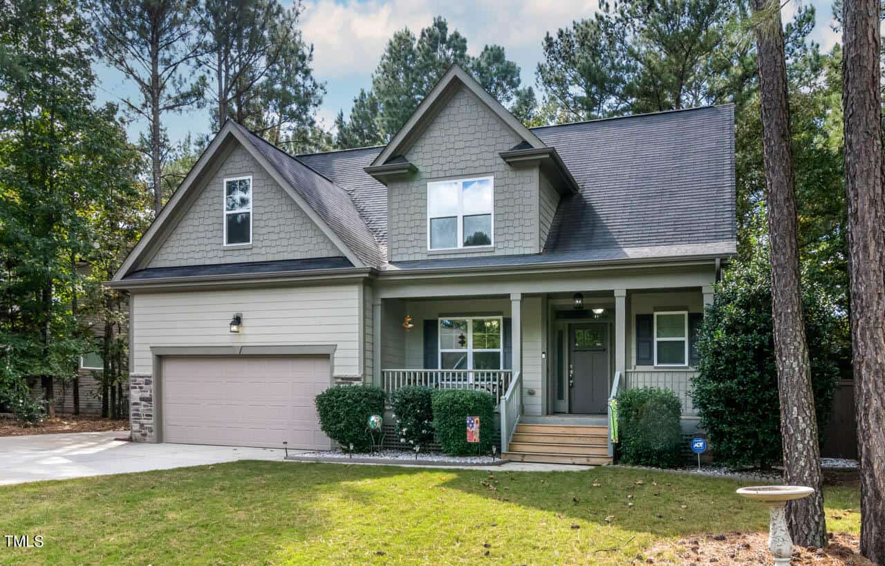 A two-story suburban house with gray siding, double garage, front porch with steps, and a well-kept lawn, surrounded by tall trees and greenery.