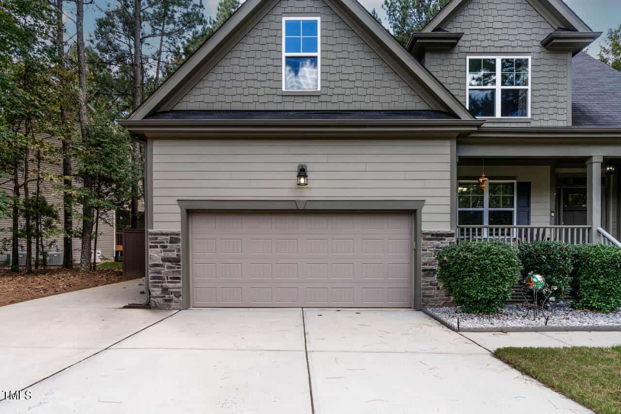 A two-car garage with a light brown door, stone accents, gray siding, and a concrete driveway; part of a modern house with a front porch, green shrubs, and trees in the background.