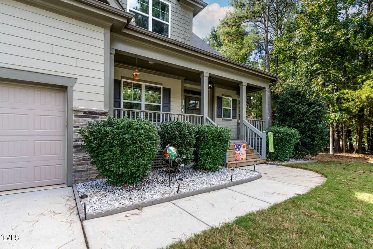 A modern house with gray siding, stone accents, and a covered front porch. Shrubs, white gravel, and lawn decor line the sidewalk leading to the porch; trees are visible in the background.