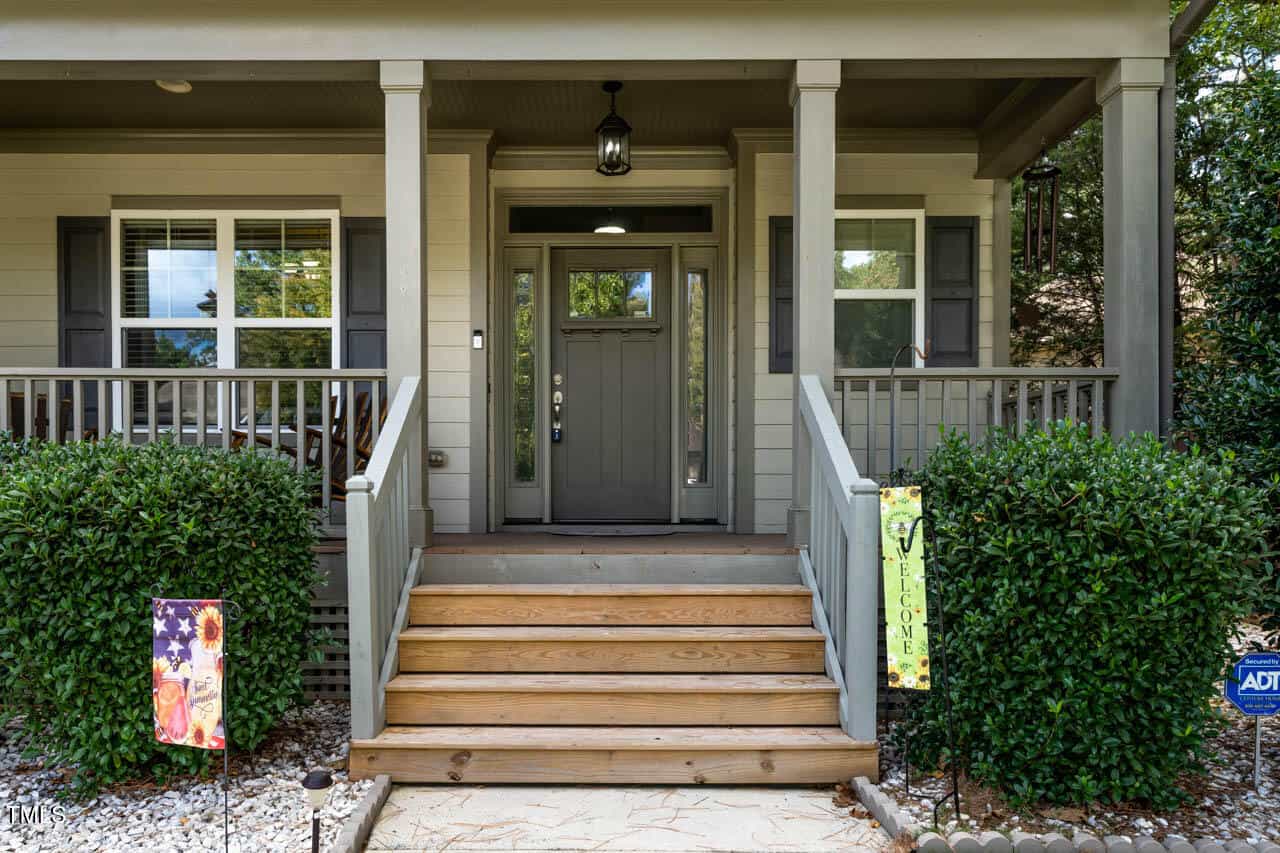 A front porch with wooden steps, gray railings, and a dark gray front door with glass panels. Shrubs border both sides, and welcome signs and a security sign are visible near the entrance.