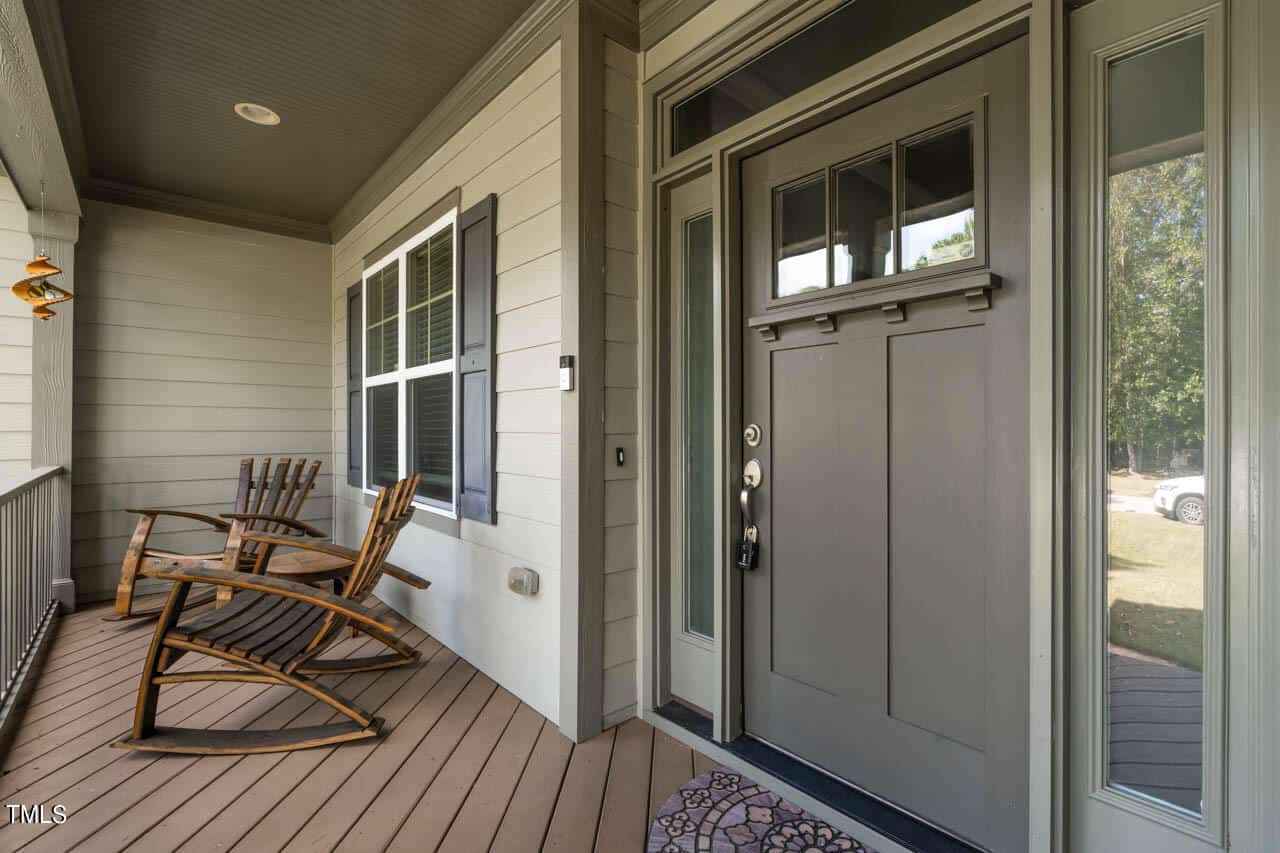 A front porch with a gray door, sidelights, two wooden rocking chairs, horizontal beige siding, and a brown wood floor. A doormat sits at the entrance, and trees are visible in the background.