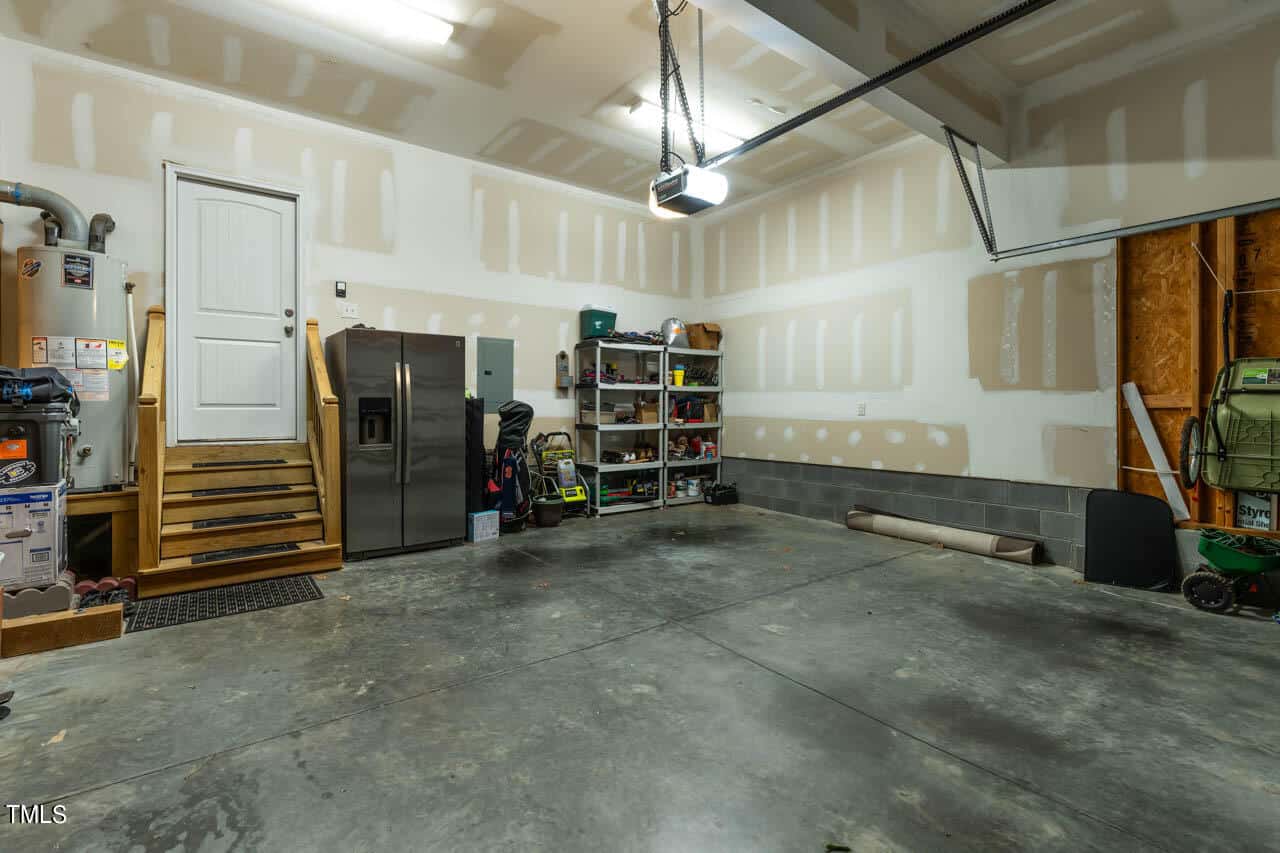 A clean, spacious garage with unfinished walls, a concrete floor, a double-door refrigerator, wooden shelves with tools and supplies, a few lawn tools, and a water heater near steps leading to a door.