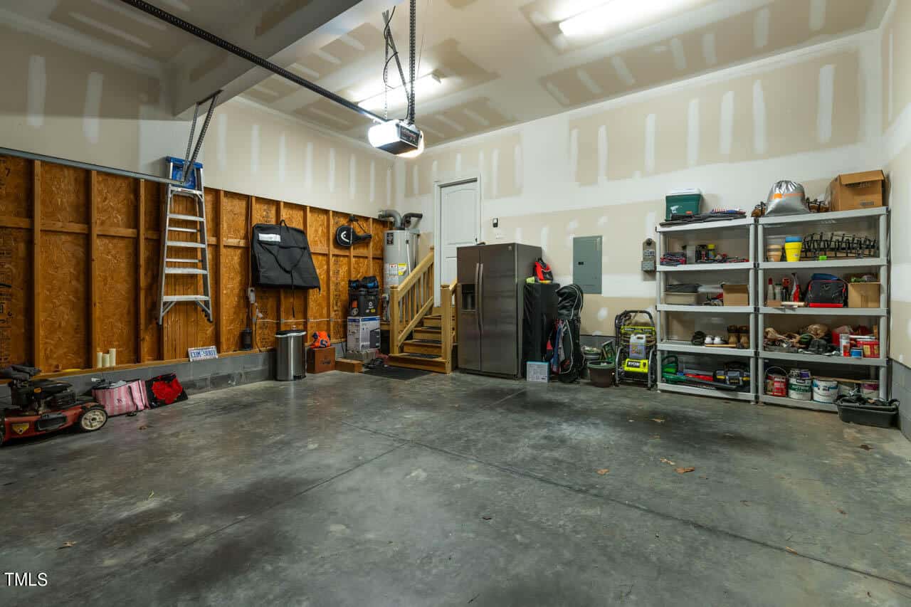 A clean, organized garage with concrete floors, unfinished walls, shelves with tools and supplies, a stainless steel refrigerator, a ladder, lawn equipment, and wooden stairs leading to a door.