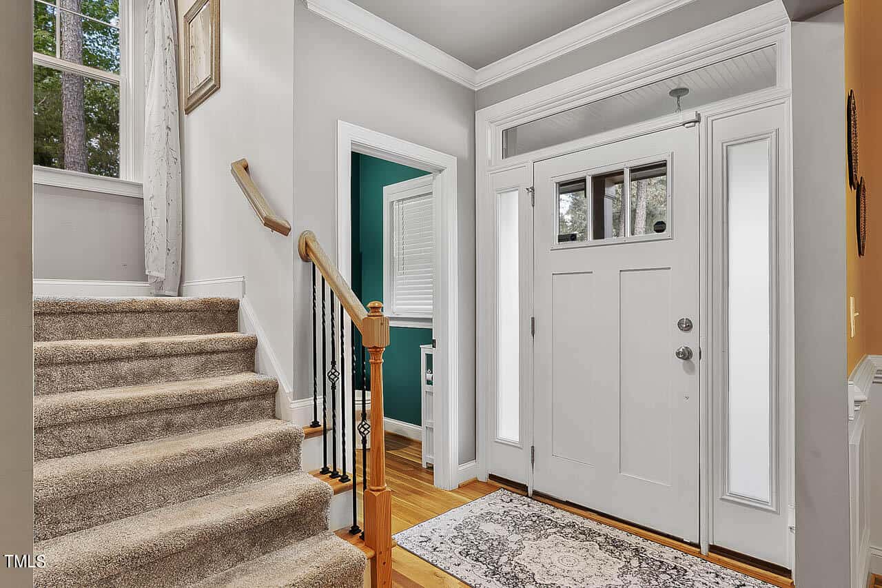 A bright entryway with a white front door, sidelights, a transom window, hardwood floor, a patterned rug, and a carpeted staircase with a wooden railing leading upstairs.