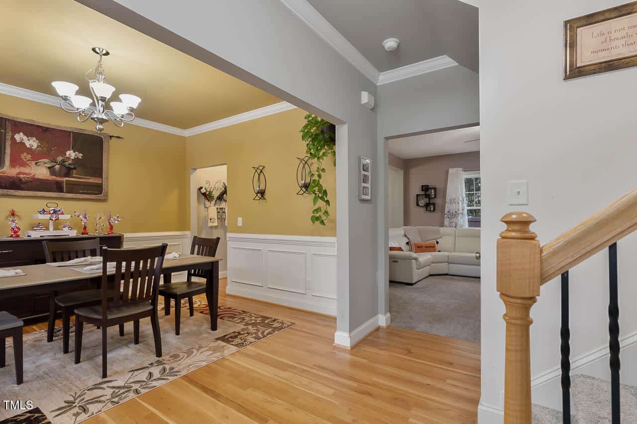 Dining room with a wooden table and six chairs, adjacent to a yellow-walled hallway and a living room with a white sofa. Light fixtures hang from the ceiling, and framed art decorates the walls. Stair railing is in the foreground.