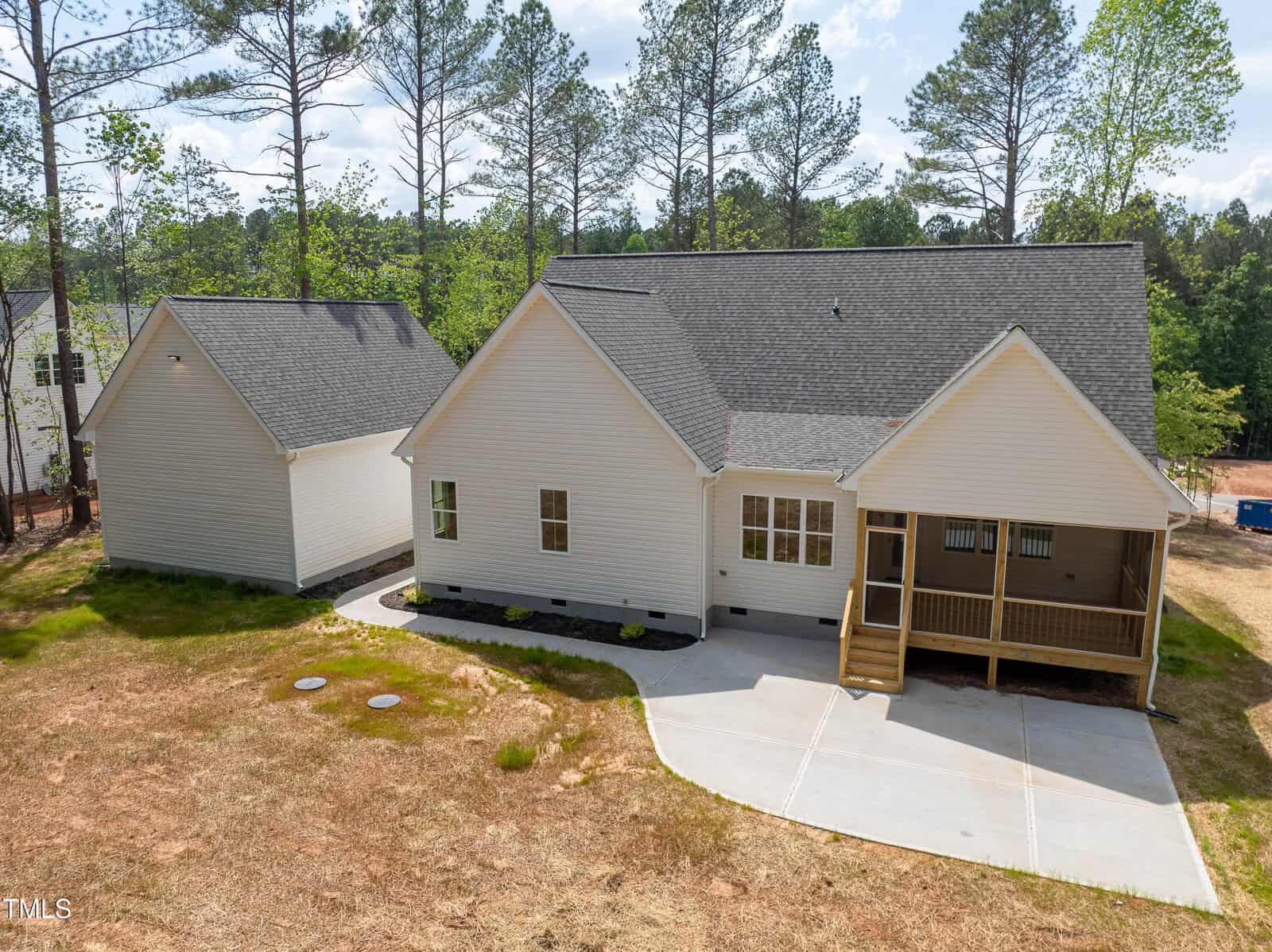 A single-story house with beige siding and dark gray shingles is shown with a detached garage, a covered back porch, and a concrete driveway, surrounded by sparse grass and trees in the background.
