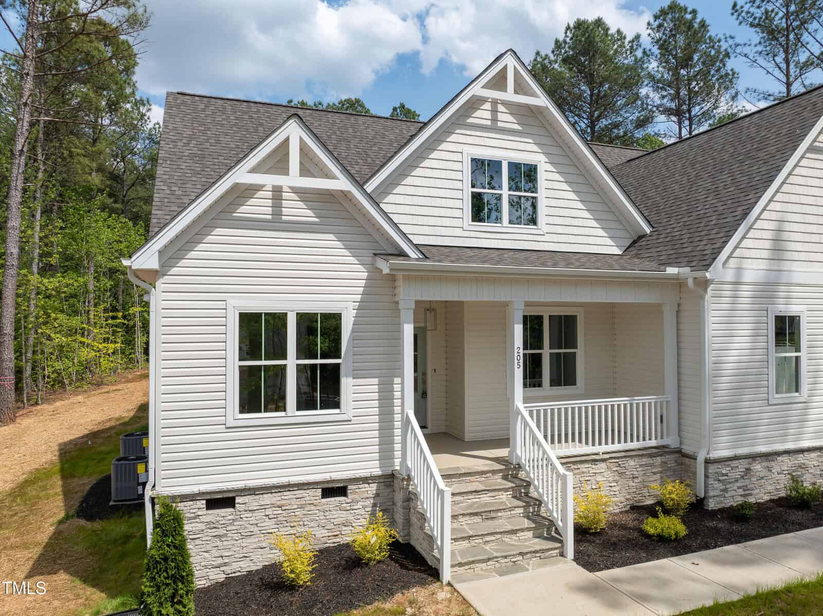 A two-story beige house with white trim, gabled roof, covered front porch, stone foundation, and landscaped yard with shrubs, surrounded by trees and greenery.