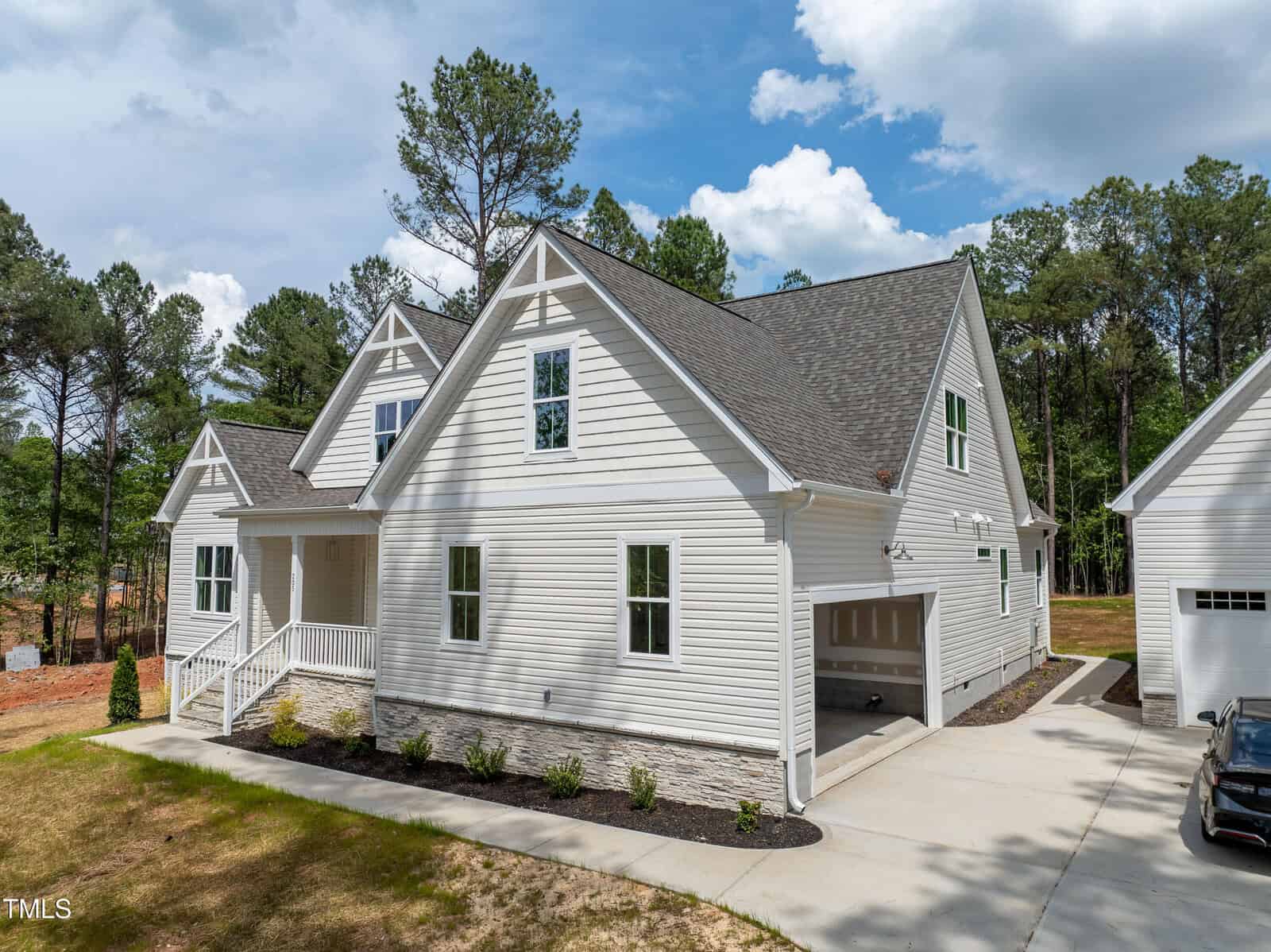 A two-story white house with gray roof shingles, attached two-car garage, front porch, and simple landscaping, set against a backdrop of tall pine trees and a blue sky with scattered clouds.