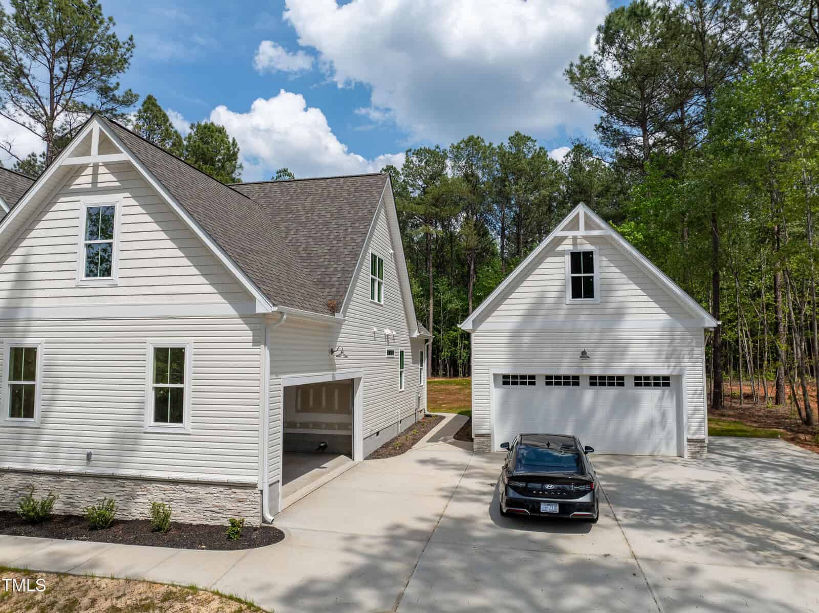A modern white house with an attached garage is shown next to a separate white two-car garage. A black car is parked in the driveway, surrounded by trees and blue sky with clouds.