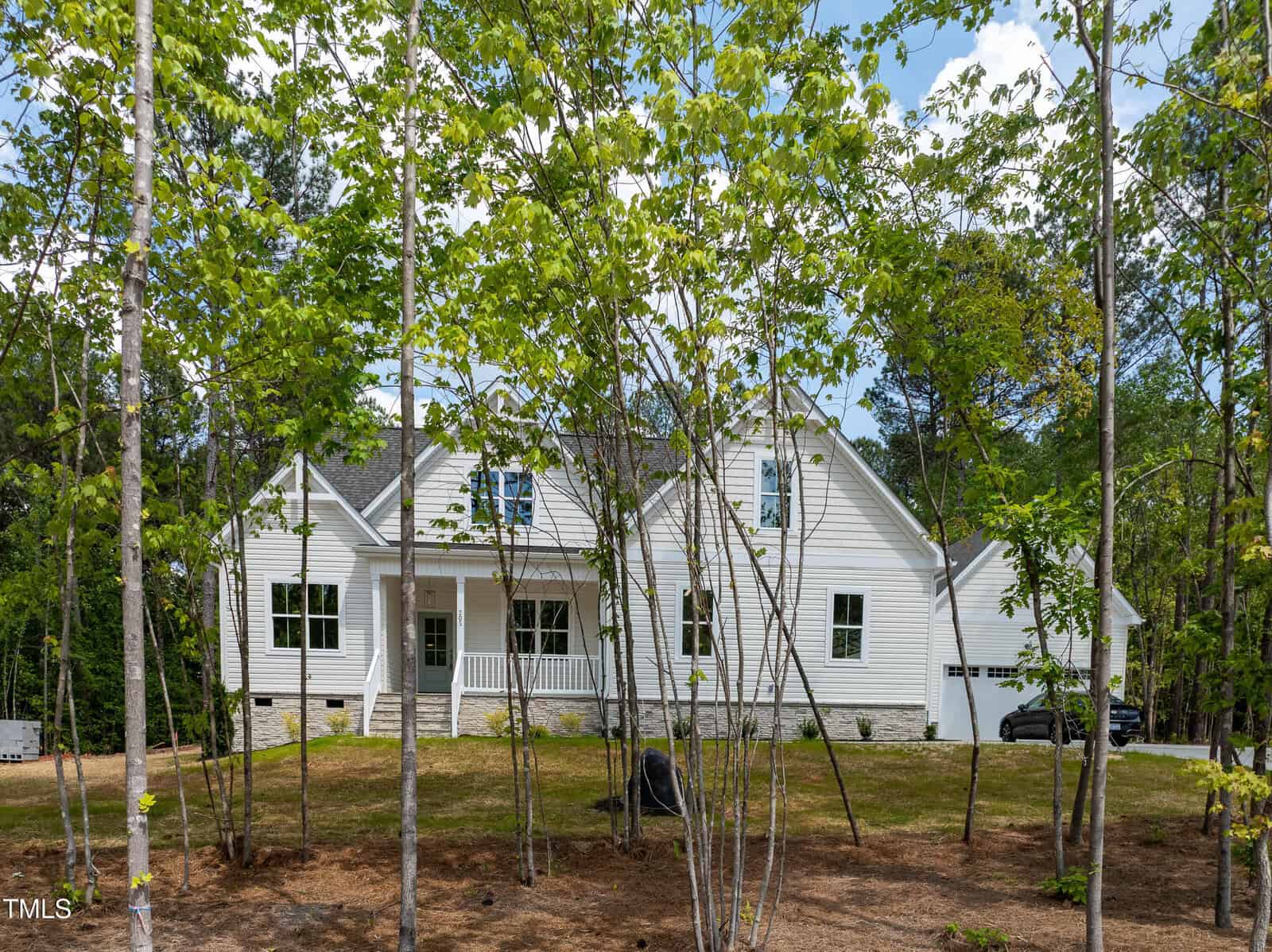 A white, two-story house with a front porch is partially obscured by young trees. The yard has sparse grass and exposed soil, and a driveway with a parked car is visible on the right side. The sky is partly cloudy.