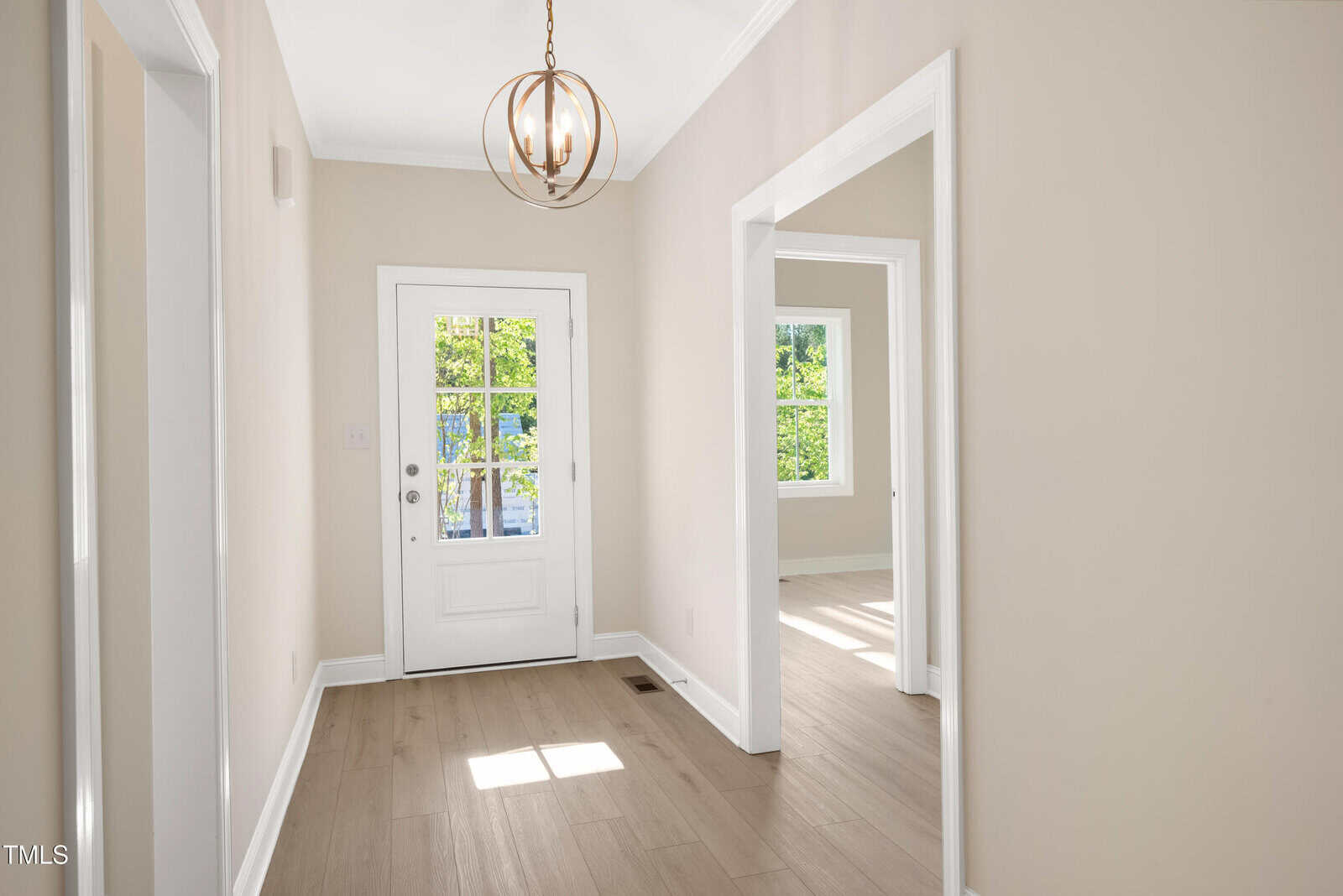 A bright hallway with light wood flooring, beige walls, white trim, a modern round light fixture, and a white front door with glass panels. Sunlight comes in through the door and nearby windows.